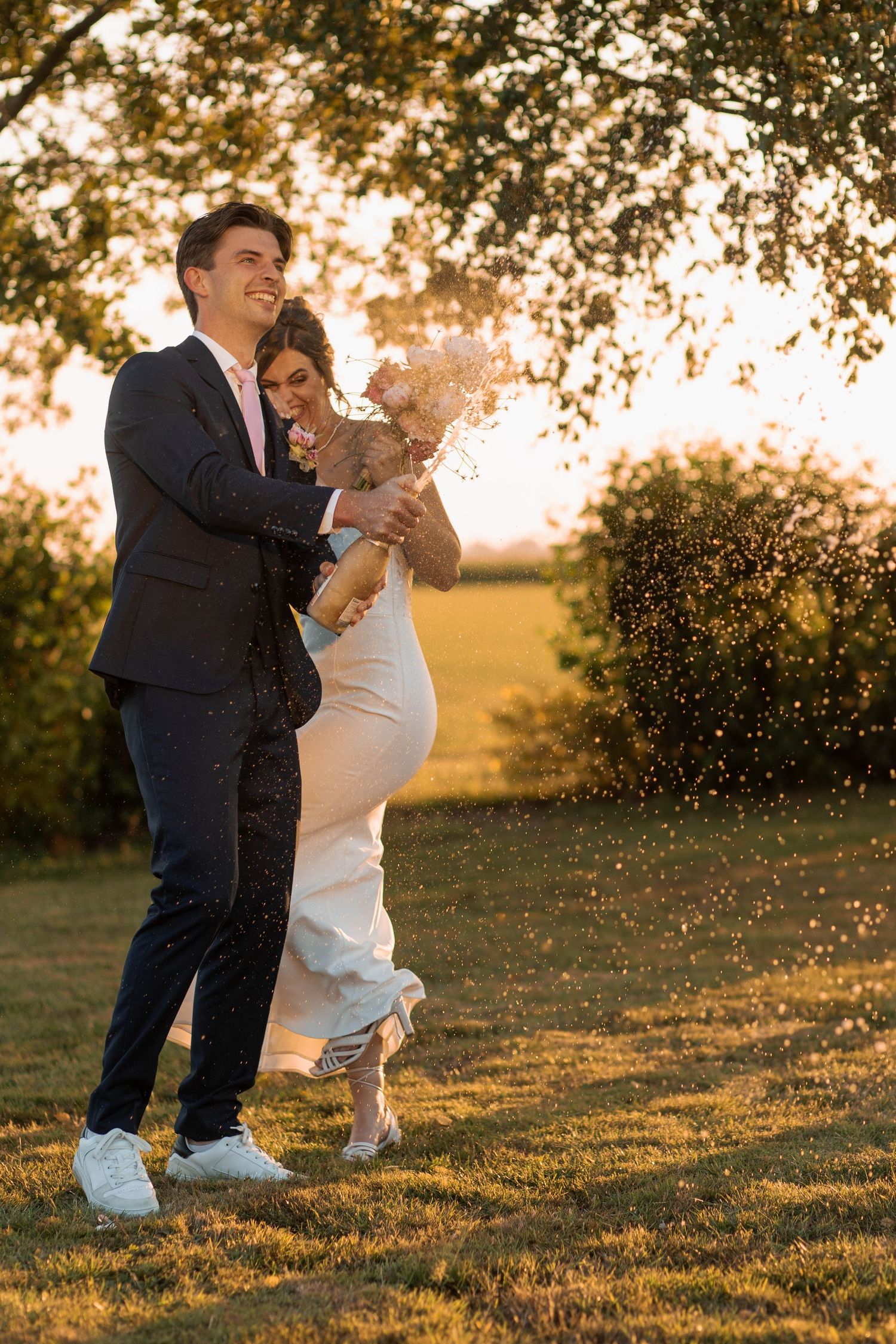 Joyful newlywed couple celebrating outdoors at golden sunset, bride in white gown holding bouquet, groom in navy suit.