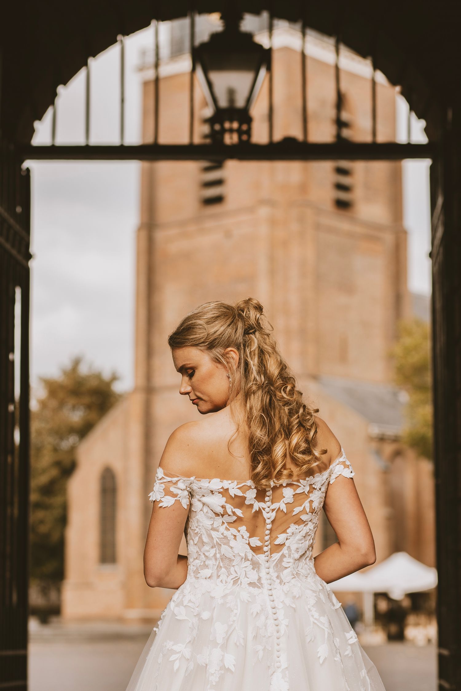 A bride in an off-shoulder lace wedding gown stands in front of a church with an arched doorway and lantern.
