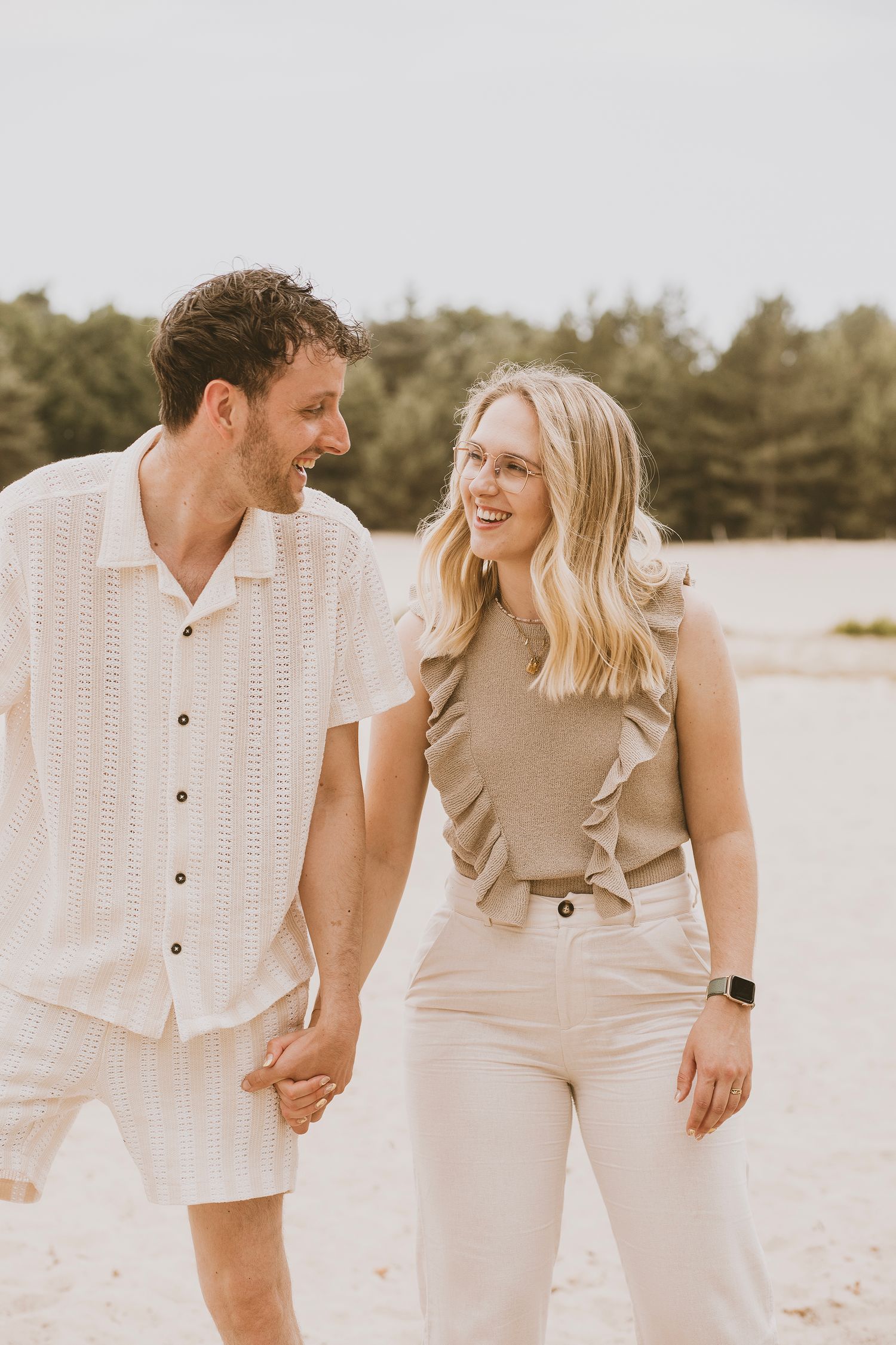 A couple in neutral beige and white outfits walks and laughs together on a sandy beach in a romantic photoshoot series.