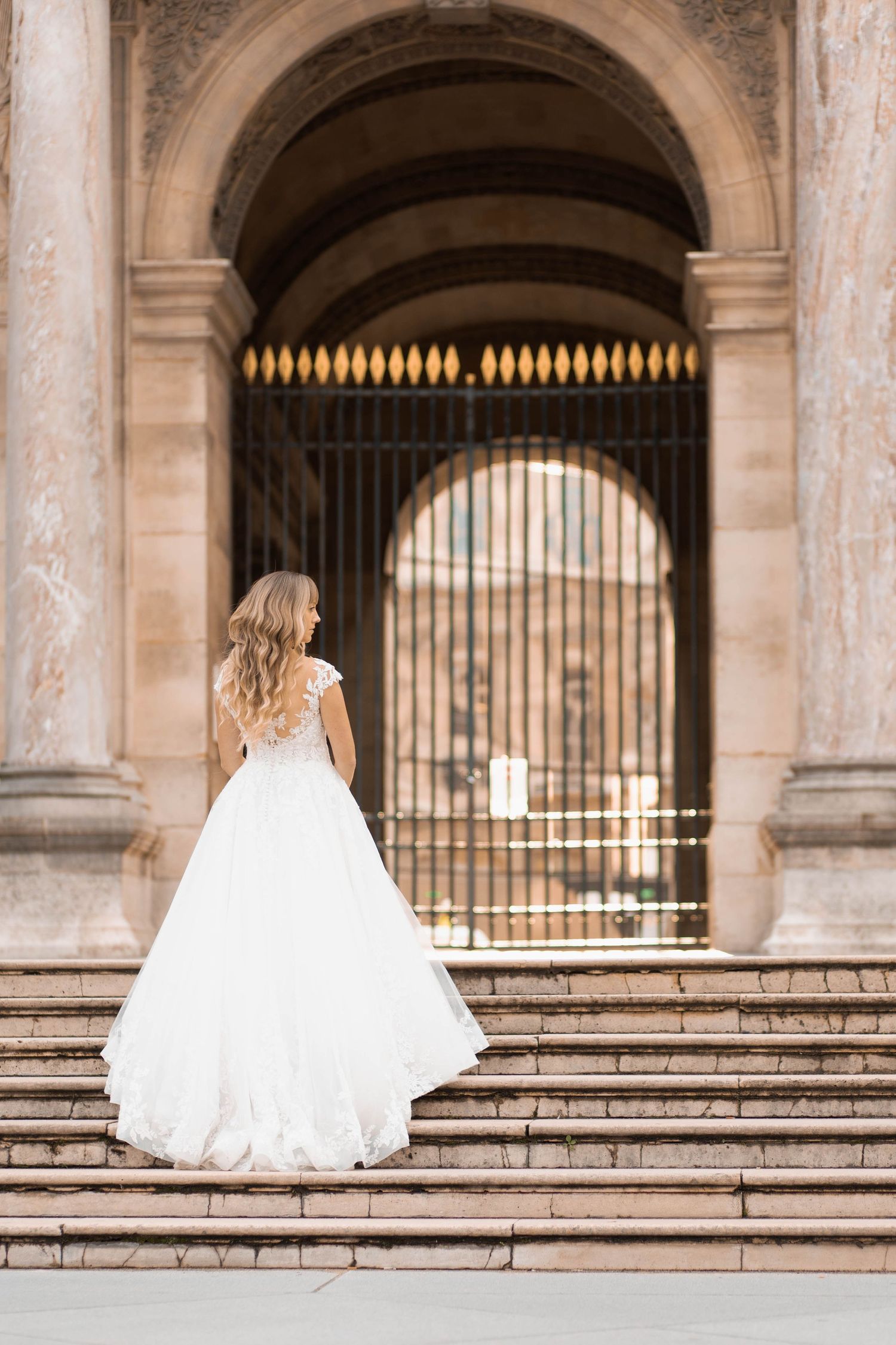 Bride in white ball gown standing on stone steps before ornate arched gate of historic building.