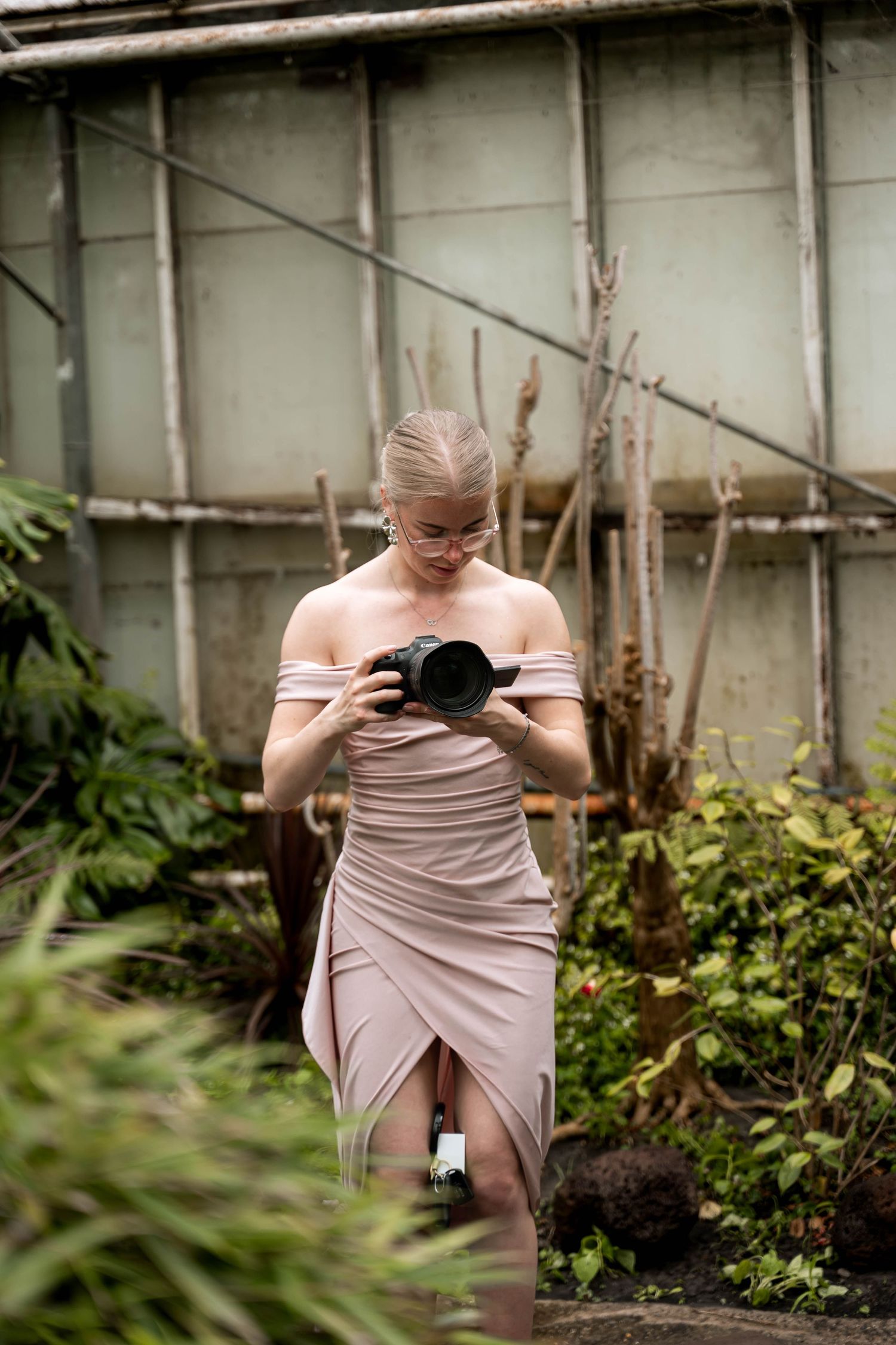 Woman in elegant blush off-shoulder dress holding DSLR camera in lush greenhouse garden setting.