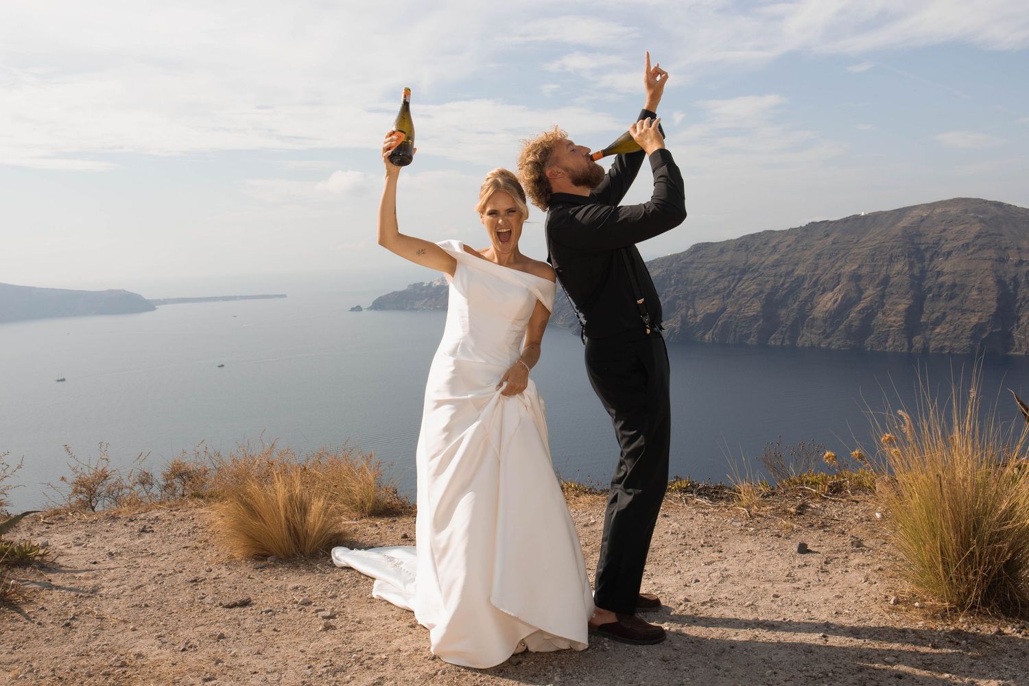 Bride in a white gown and groom in a black suit raise champagne bottles on a cliff overlooking the sea during a destination wedding.