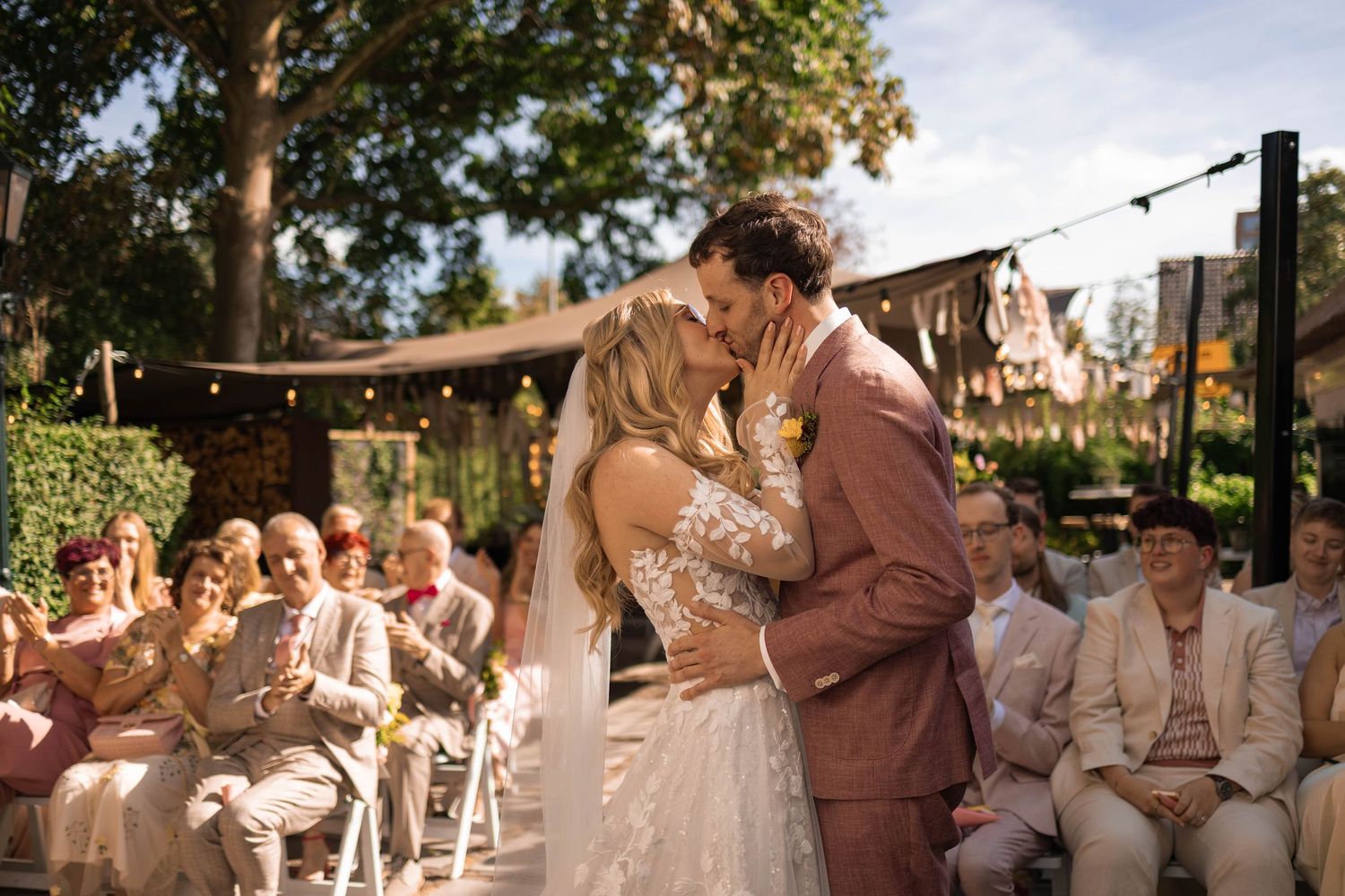 Bride and groom share a romantic kiss during an outdoor garden wedding ceremony while smiling guests look on from white chairs.