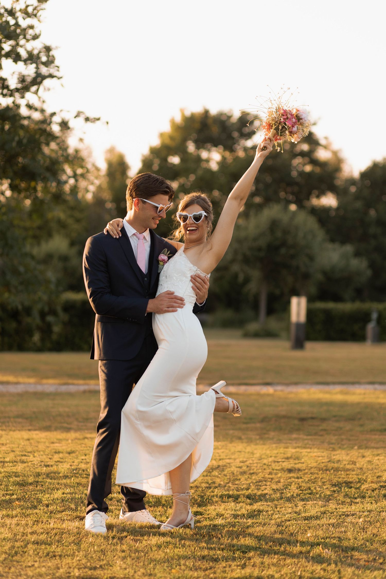 Joyful newlyweds in sunglasses celebrate outdoors at sunset, bride raising bouquet in white dress, groom in navy suit.