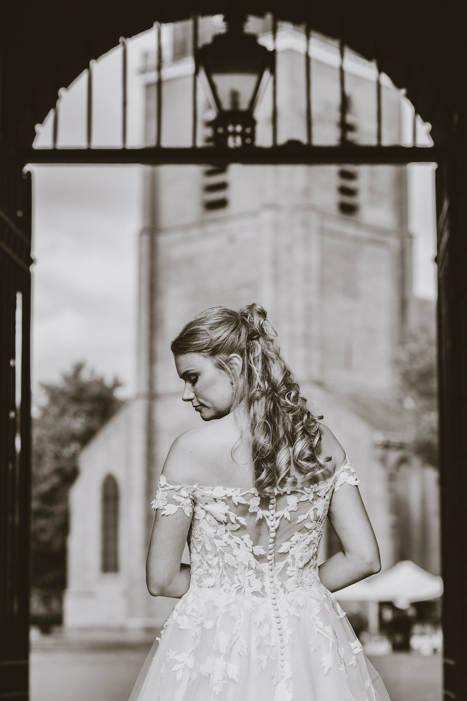 Black and white portrait of bride in lace dress against architectural backdrop with arched windows.