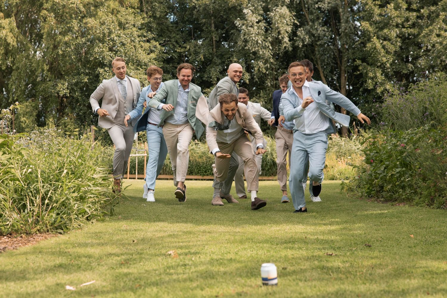 Groomsmen in matching sage shirts and tan suits racing across green lawn at outdoor wedding celebration.