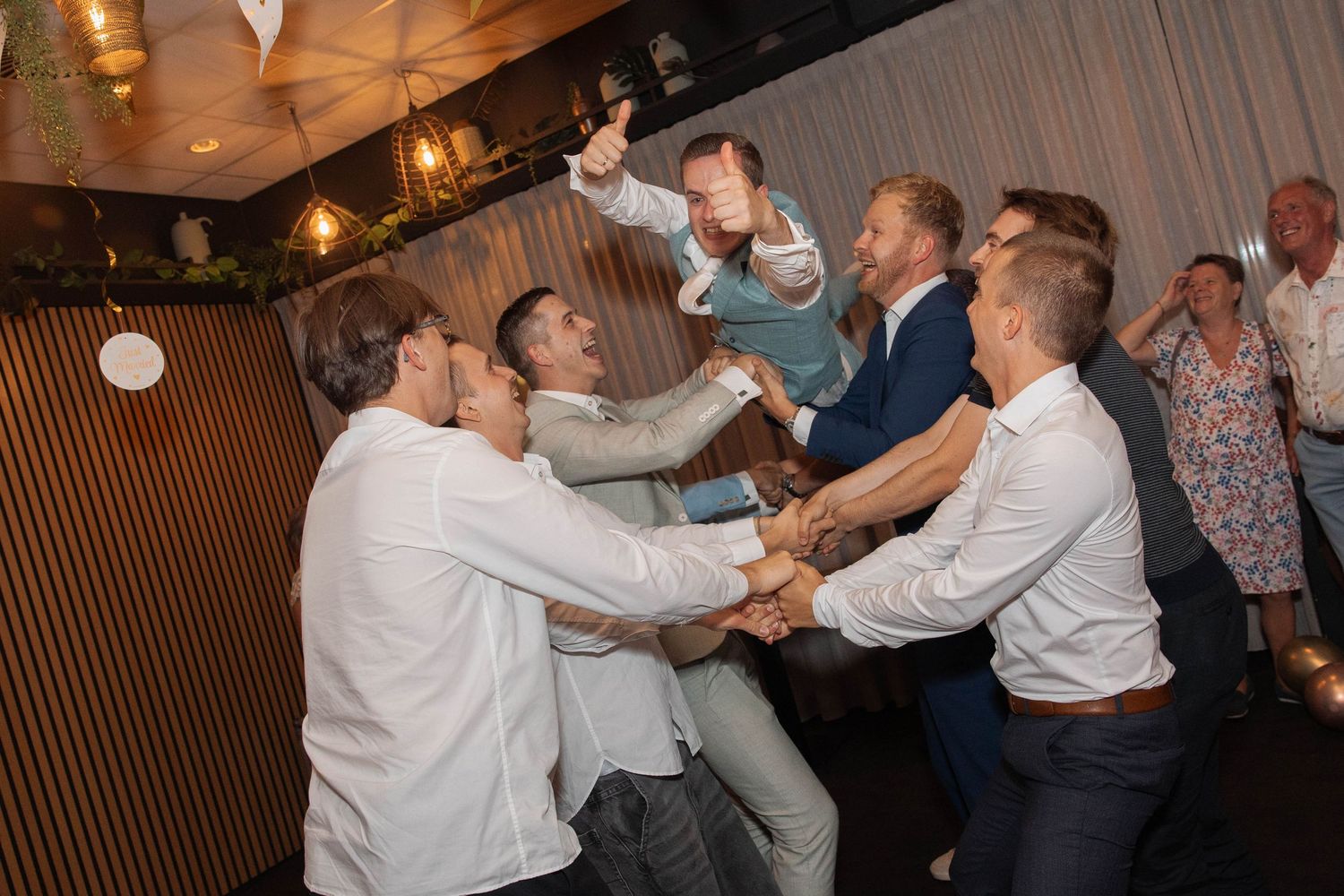 Groomsmen lifting groom in celebration at a wedding reception in a warmly lit venue.