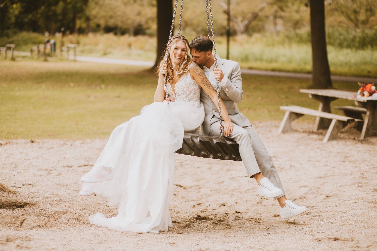 A couple in wedding attire sits together on a wooden swing in a rustic outdoor setting during golden hour.