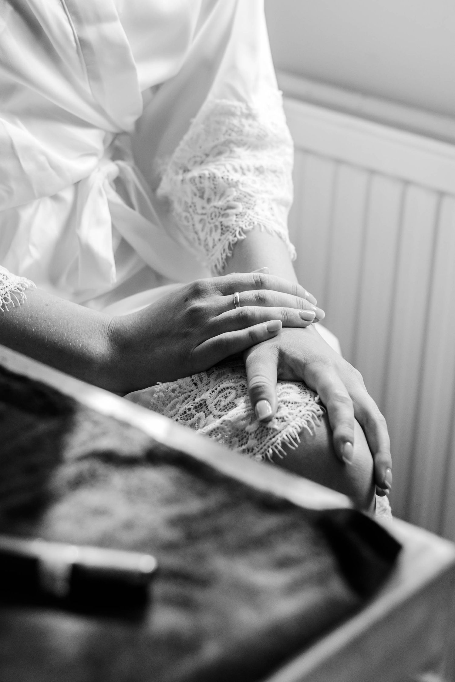 Black and white photo of a bride's hands resting gently, wearing an engagement ring, with a lace dress and white robe.