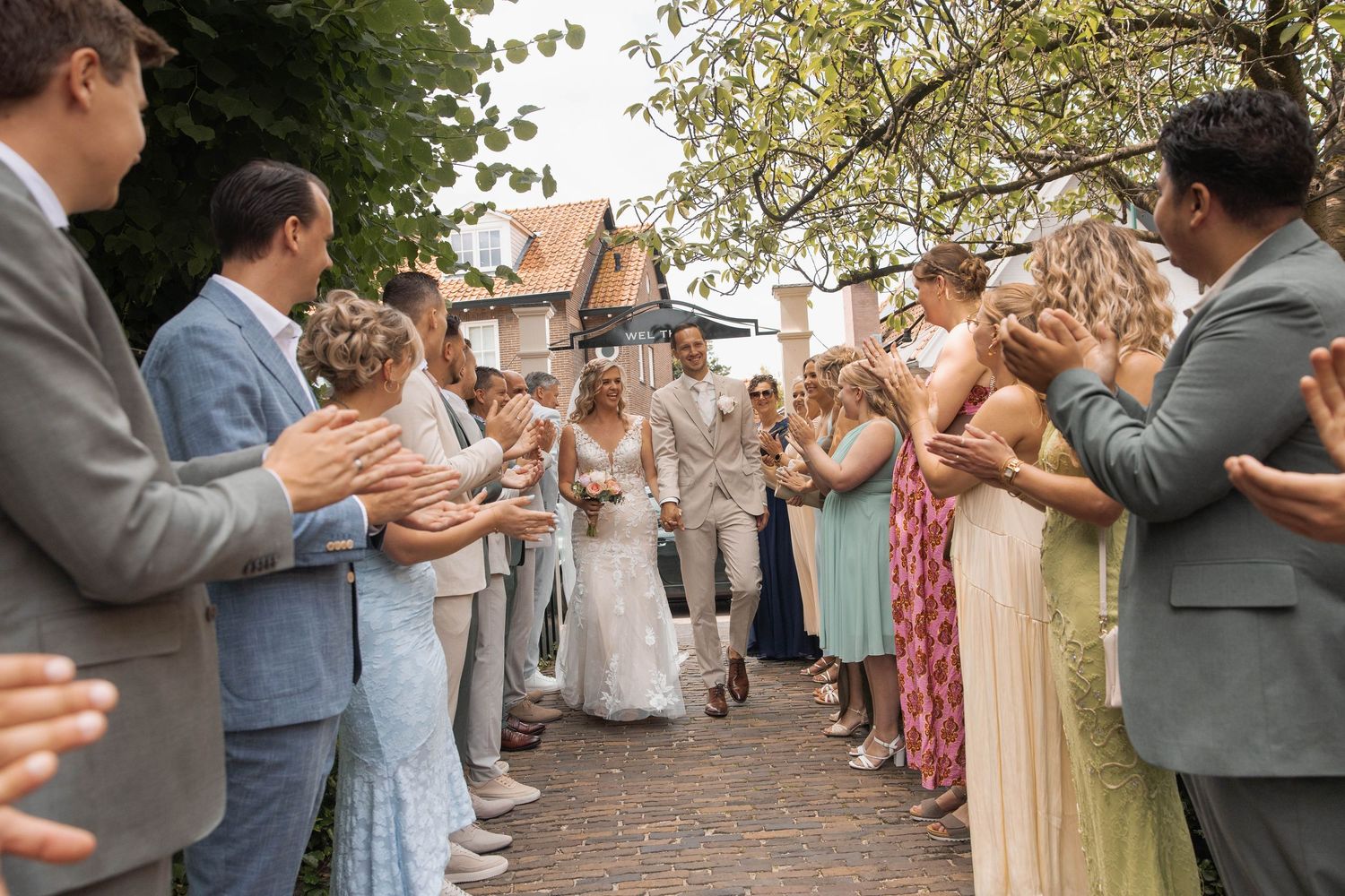 Bride and groom walk through applauding wedding guests outdoors at a charming venue surrounded by trees.