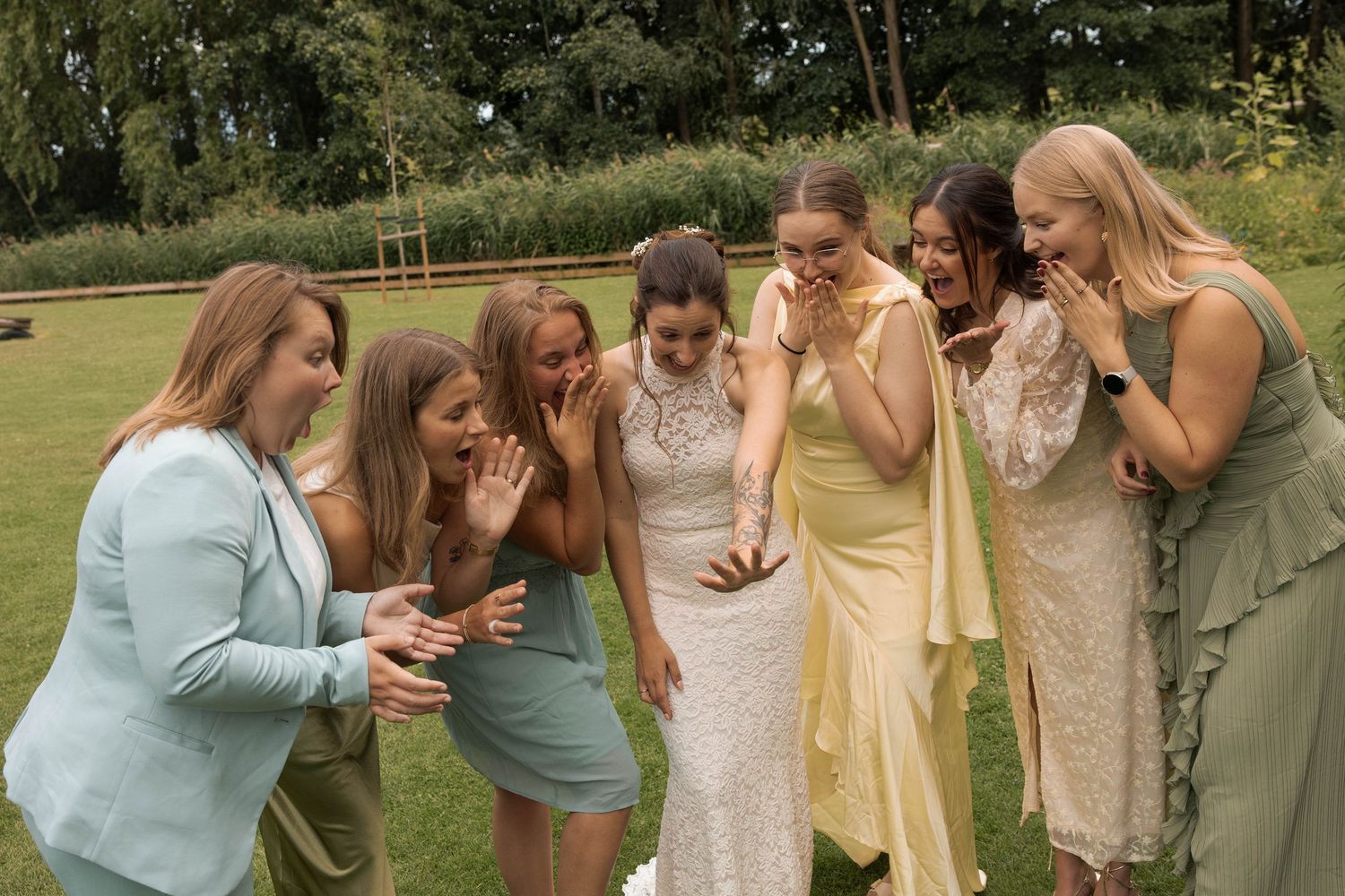 Bride surrounded by joyful bridesmaids in green and yellow dresses reacting with excitement outdoors at a wedding.