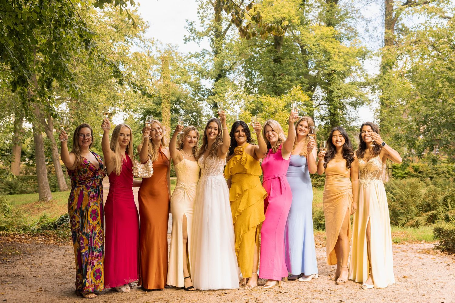 Group of bridesmaids in colorful dresses posing outdoors in a garden setting with autumn trees.