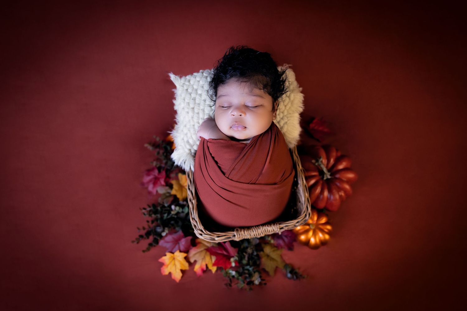 Newborn baby swaddled in rust wrap, sleeping in basket with autumn flowers on red background.