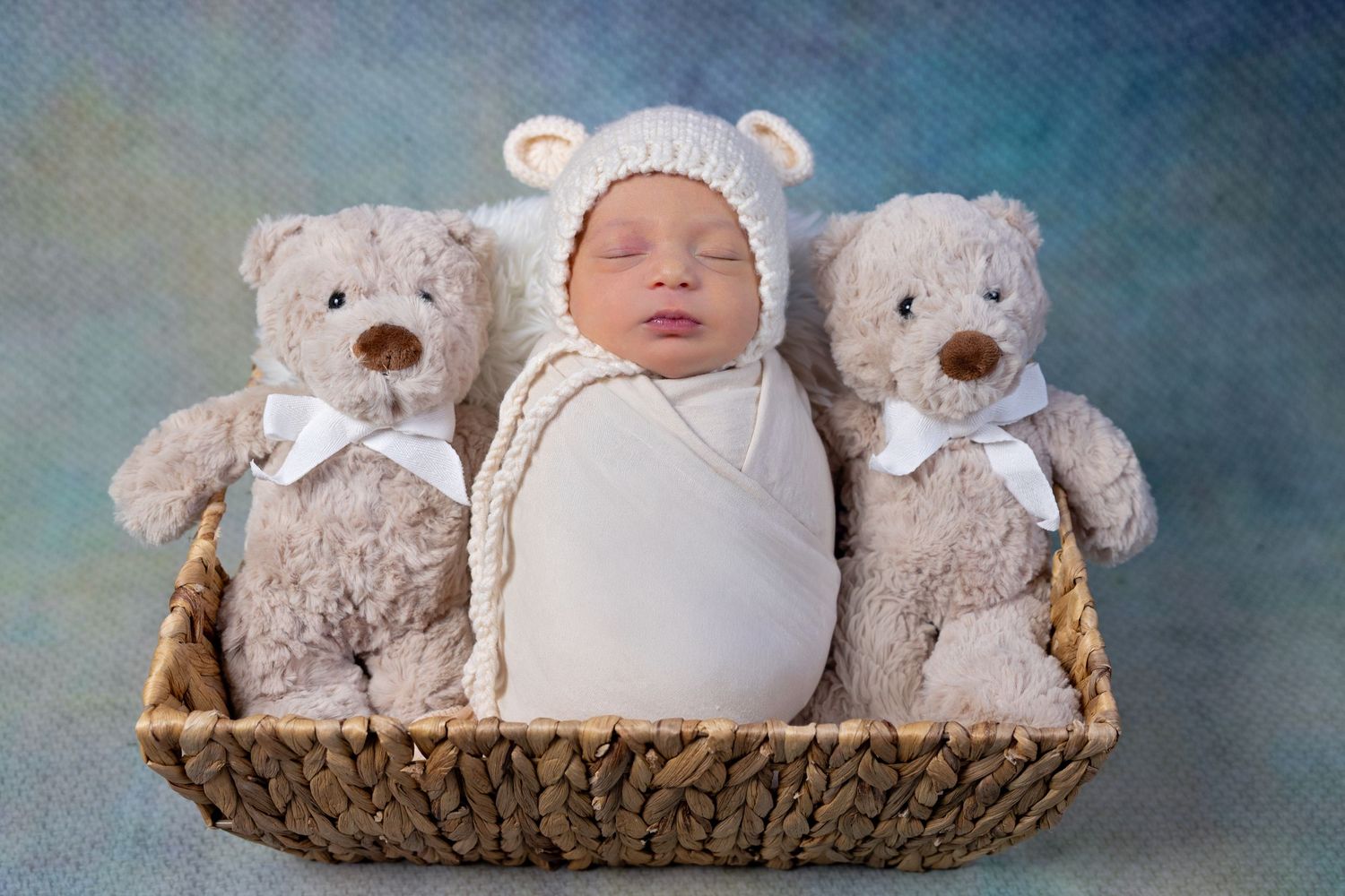 Newborn baby swaddled in white, wearing a bear hat, nestled between two teddy bears in a wicker basket.