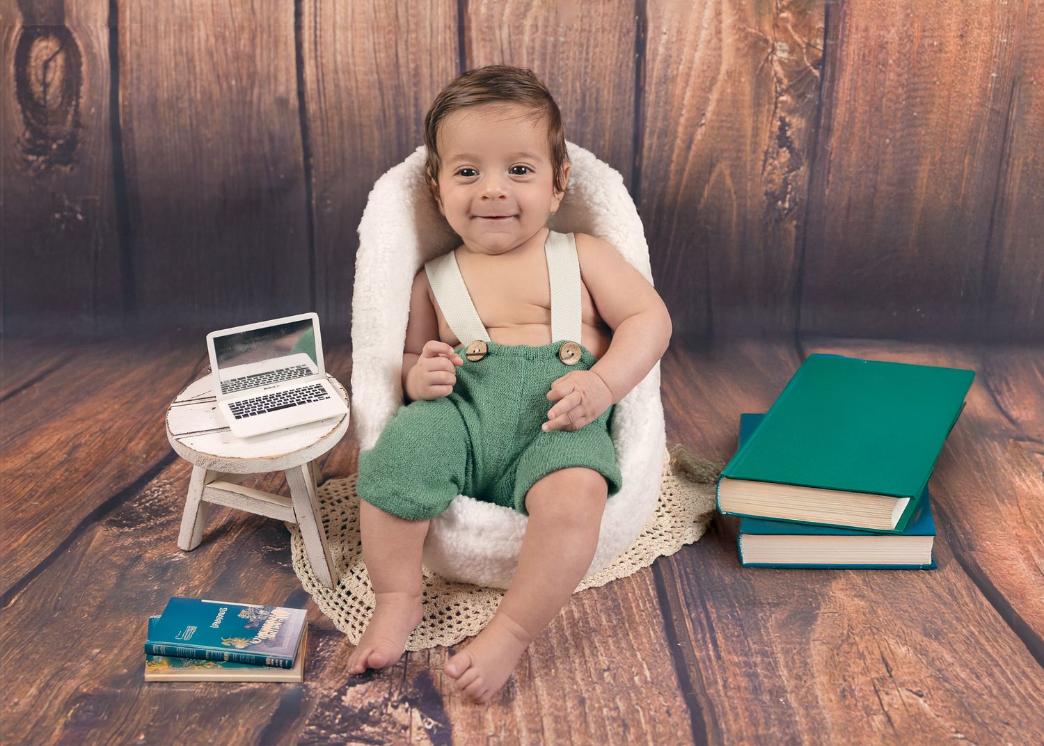 Smiling baby in green overalls sits in white chair with books and toy laptop on wooden floor.