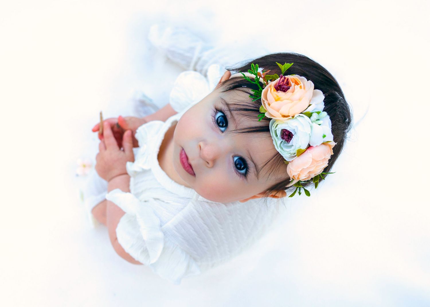 Adorable baby girl with big blue eyes wearing a white outfit and floral headband with pink and white roses.
