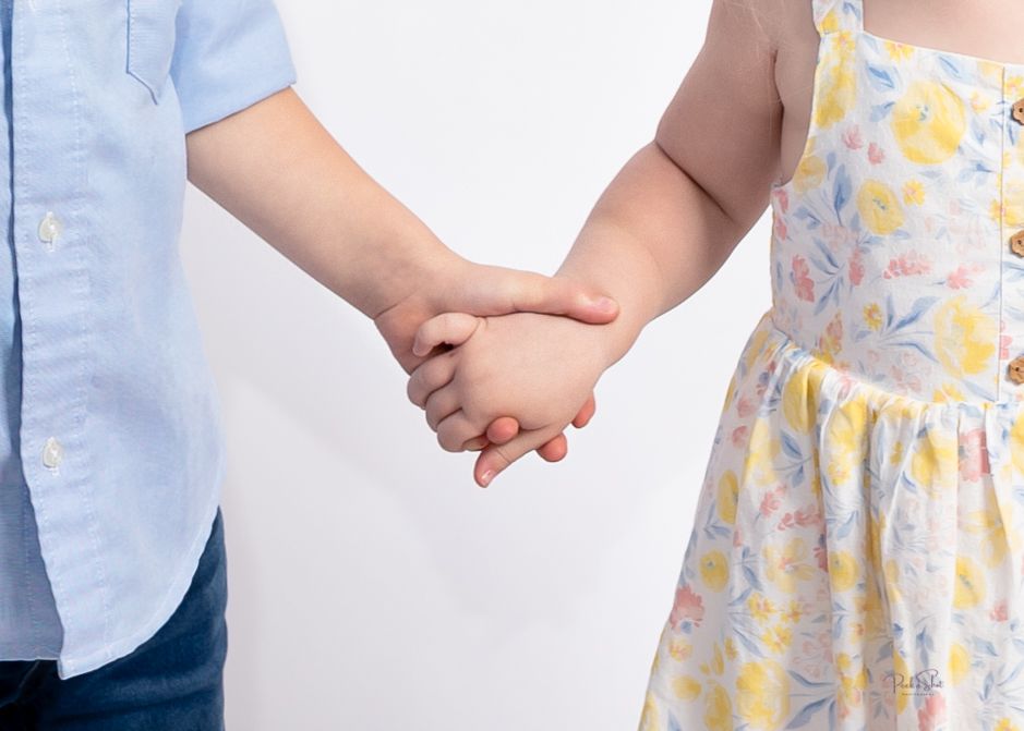 Two young children holding hands, a boy in a blue shirt and a girl in a floral dress, against a white background.