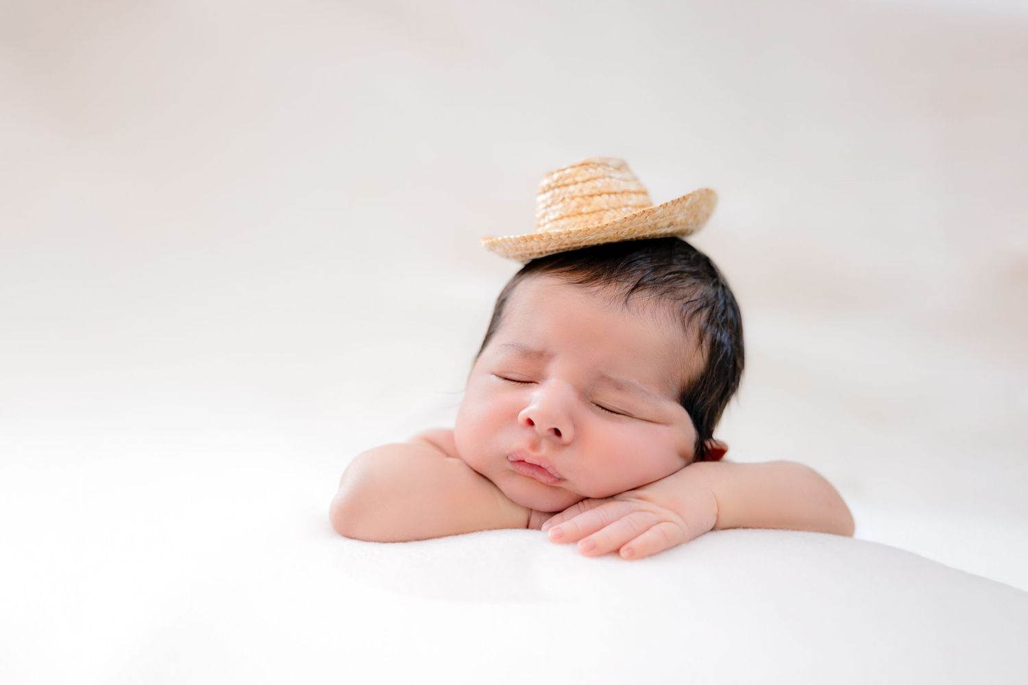 Sleeping newborn baby wearing a tiny straw hat, resting chin on hands against a white background.