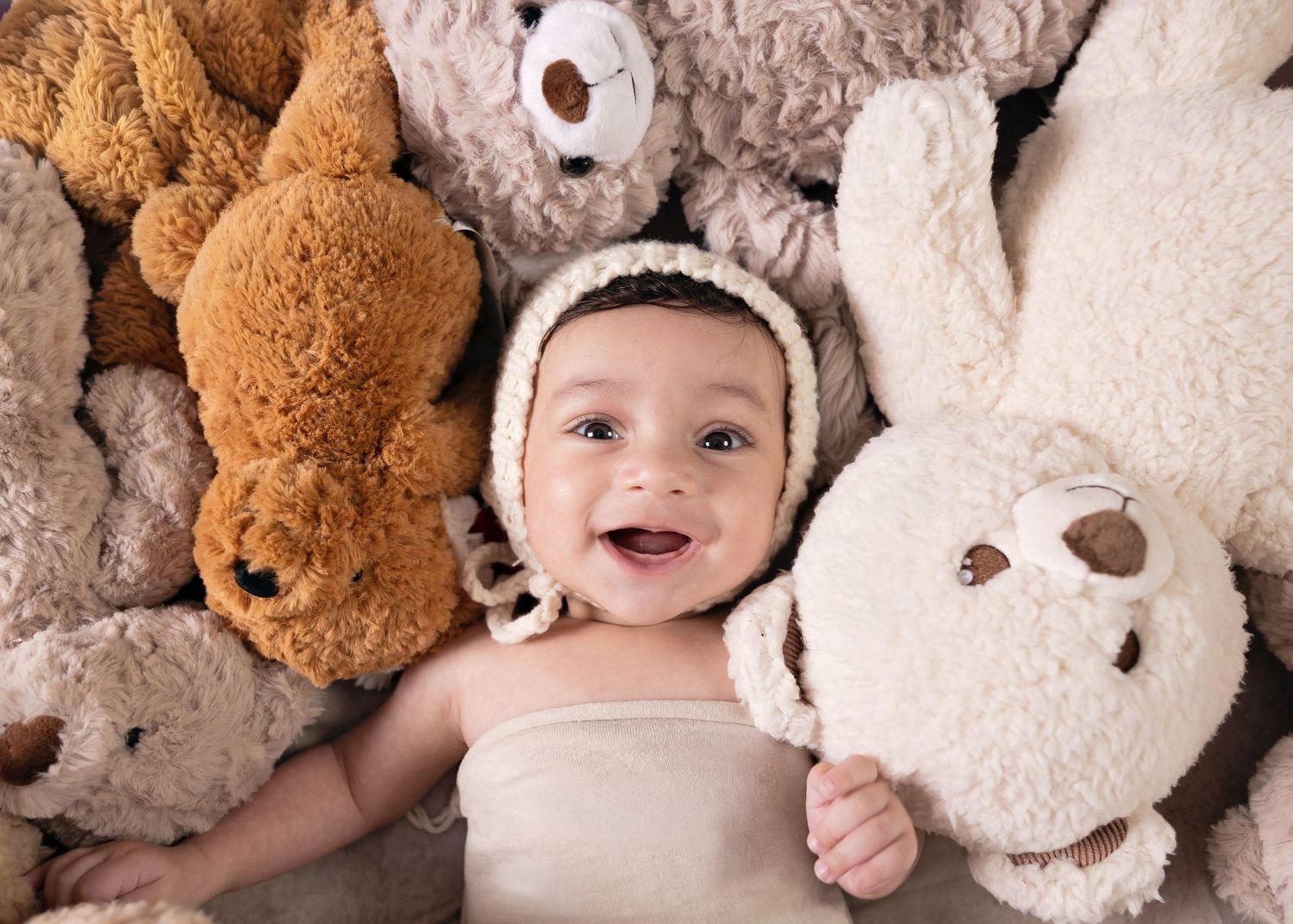 Smiling baby in knit bonnet surrounded by soft plush teddy bears and bunny stuffed animals in neutral tones.
