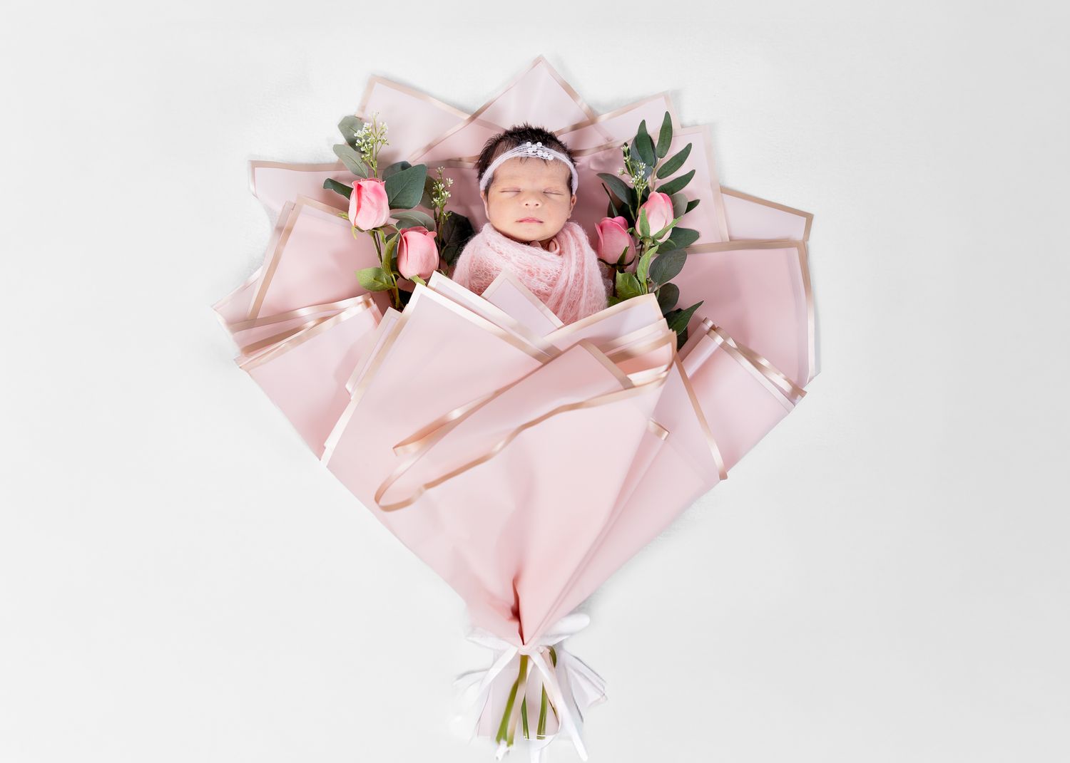 Newborn baby girl wrapped in pink swaddle, posed as a flower bouquet with pink roses and greenery on white background.