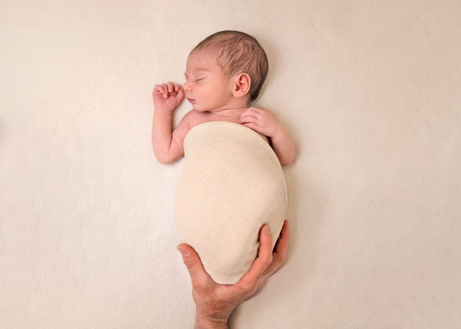 Newborn baby swaddled in cream wrap, sleeping peacefully while adult hands gently support them on a soft beige background.