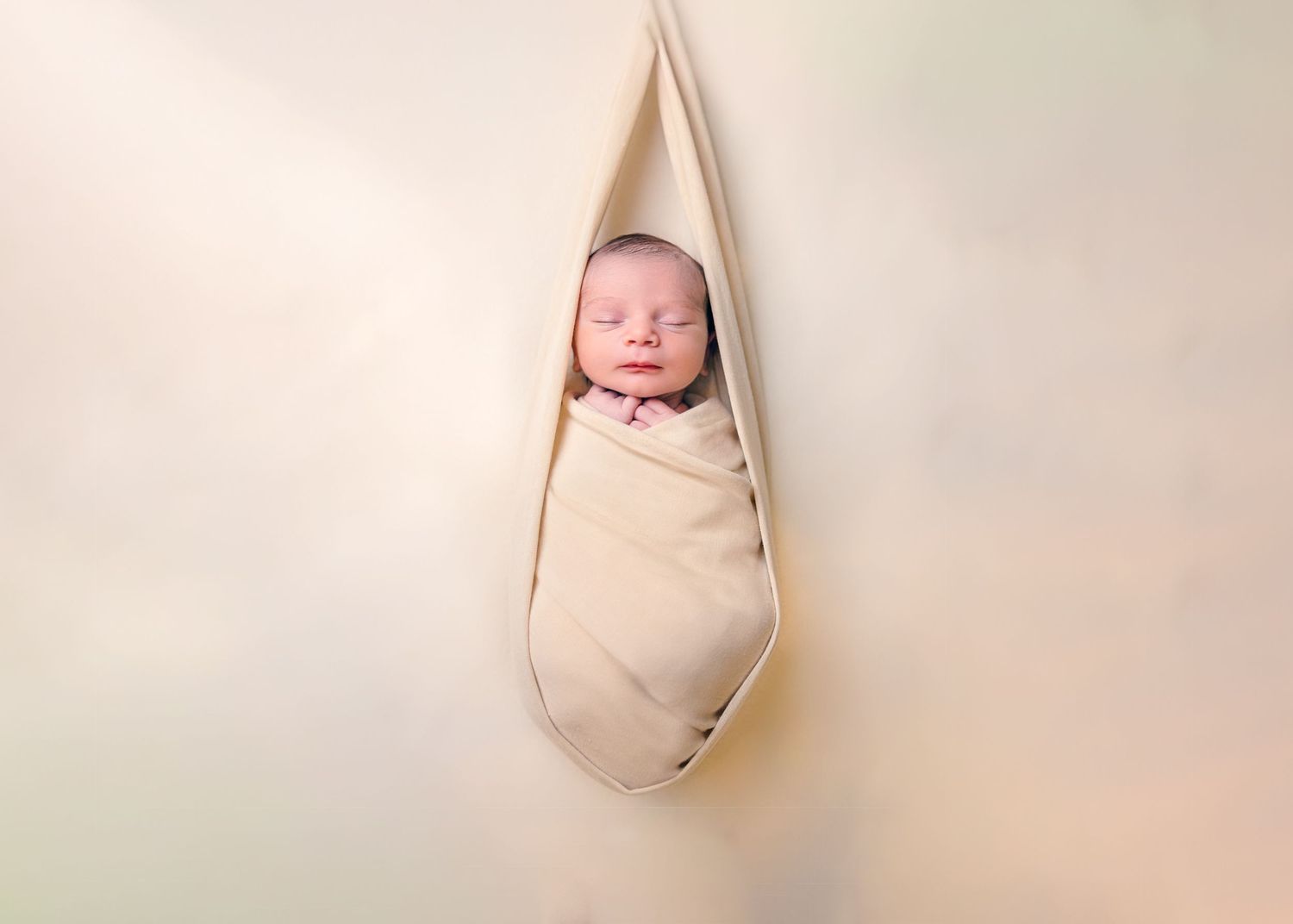 Newborn baby swaddled in cream wrap, peacefully sleeping in a hanging hammock against a soft beige background.