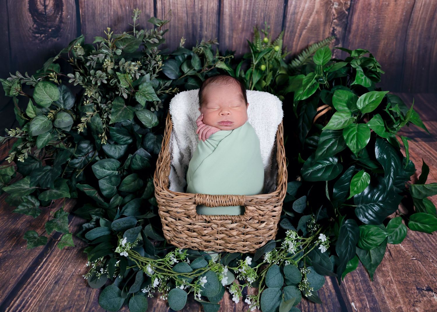 Newborn baby swaddled in green wrap sleeping in wicker basket surrounded by lush green foliage on wood backdrop.