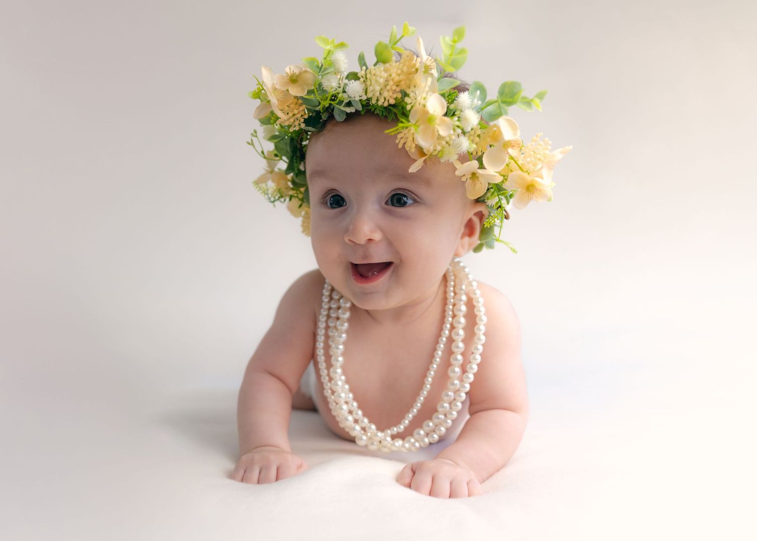 Smiling baby wearing a floral crown and pearl necklace poses on a white background.