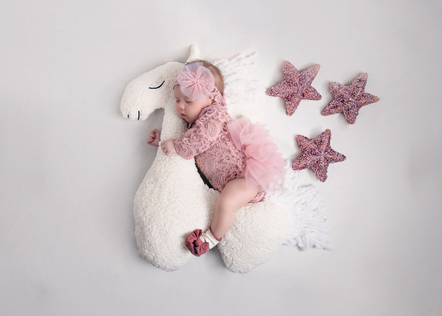 Newborn baby in pink tutu sleeping on white unicorn plush with crocheted pink stars on white background.