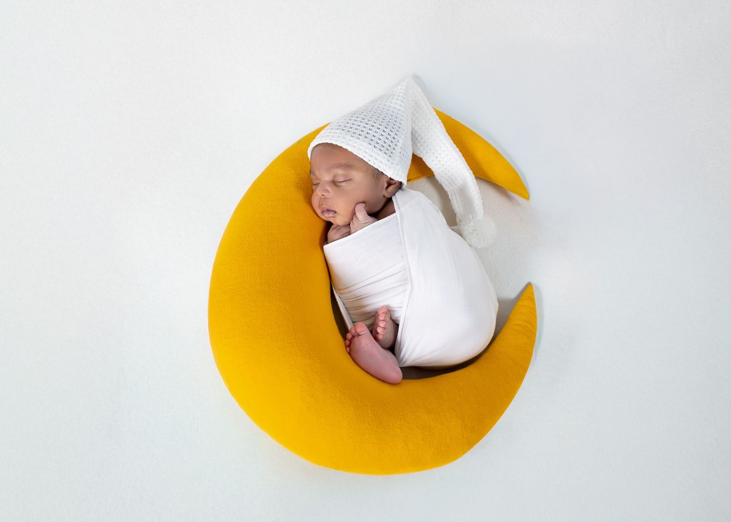 Newborn baby swaddled in white wrap, sleeping peacefully in a yellow moon-shaped prop with a white bunny ear hat.
