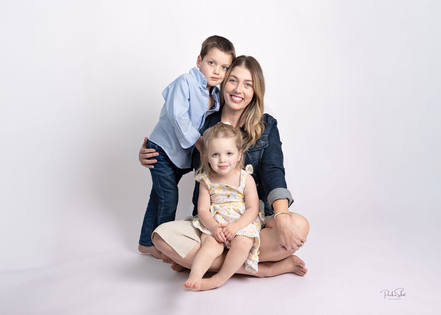 A smiling mother sits cross-legged with her toddler daughter on her lap and young son hugging her from behind.