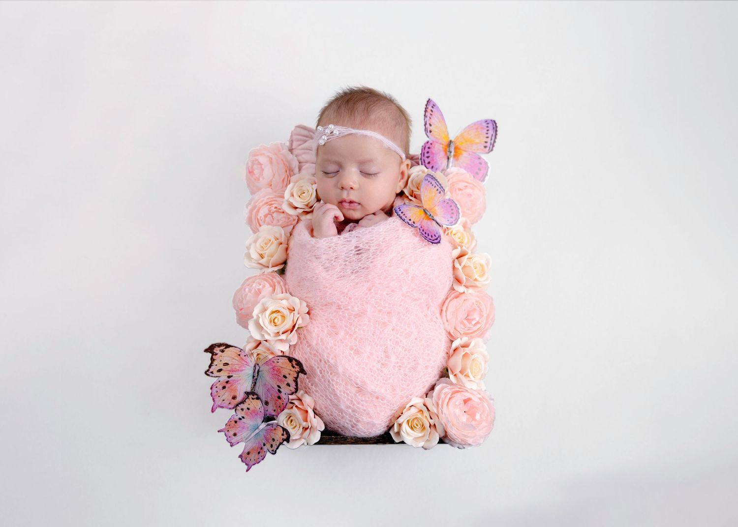 Sleeping newborn baby wrapped in pink swaddled in floral frame with colorful butterflies on white background.