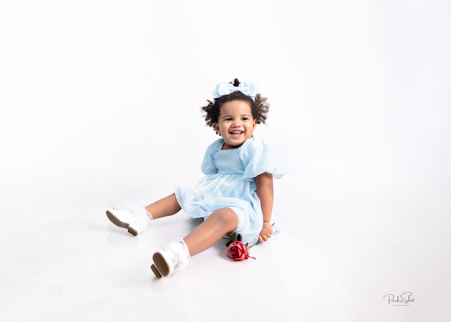 Smiling toddler girl in light blue dress sitting on white background, wearing white shoes and hair accessories.