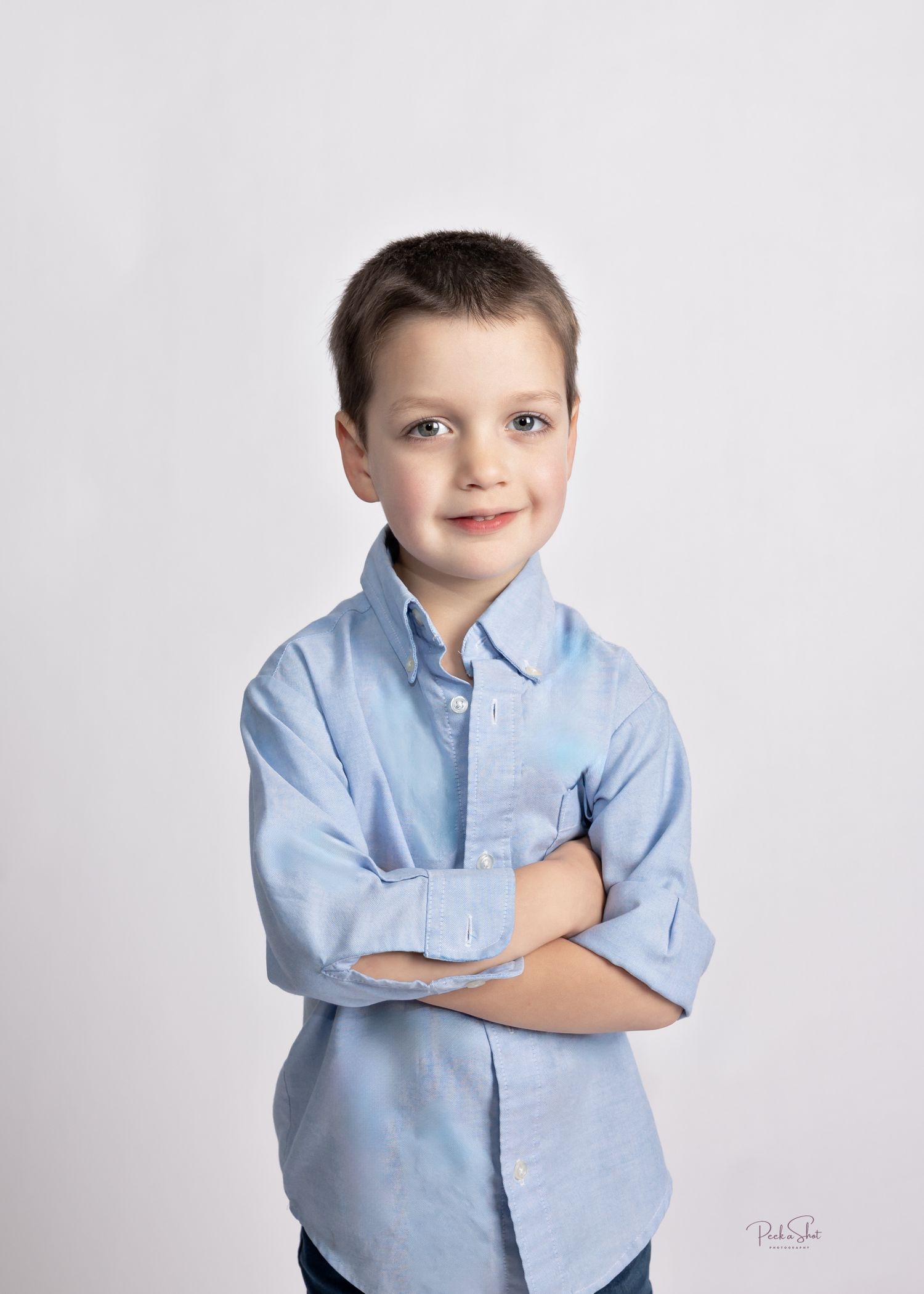 Young boy in a light blue button-up shirt poses with arms crossed, smiling confidently against a white background.