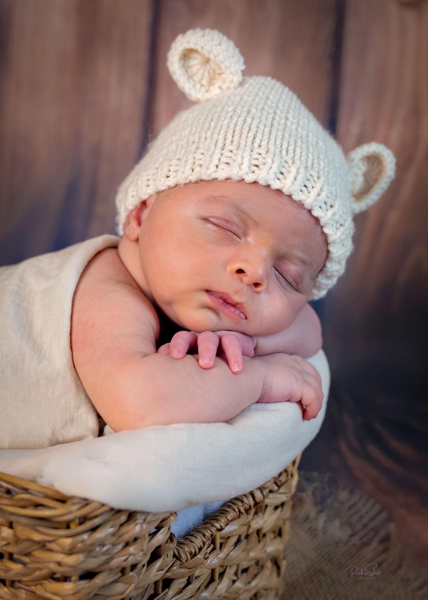 Sleeping newborn baby in a wicker basket wearing a cream knit bear ear hat, wrapped in soft white fabric.