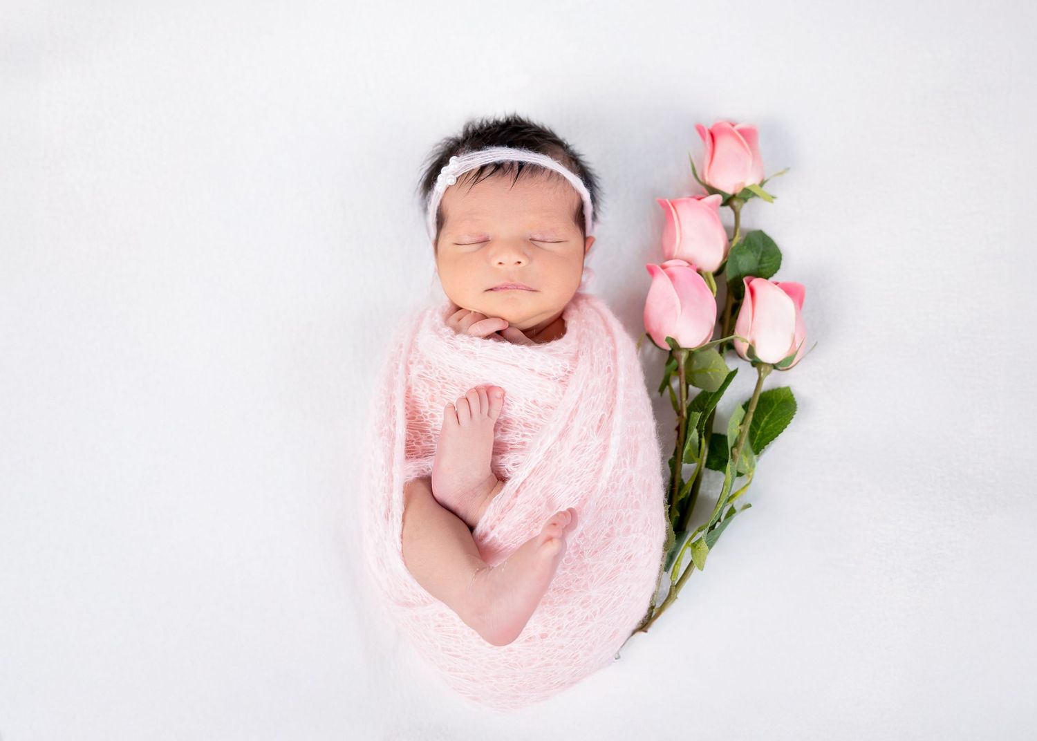 Newborn baby swaddled in pink wrap sleeping beside pink roses on white background.