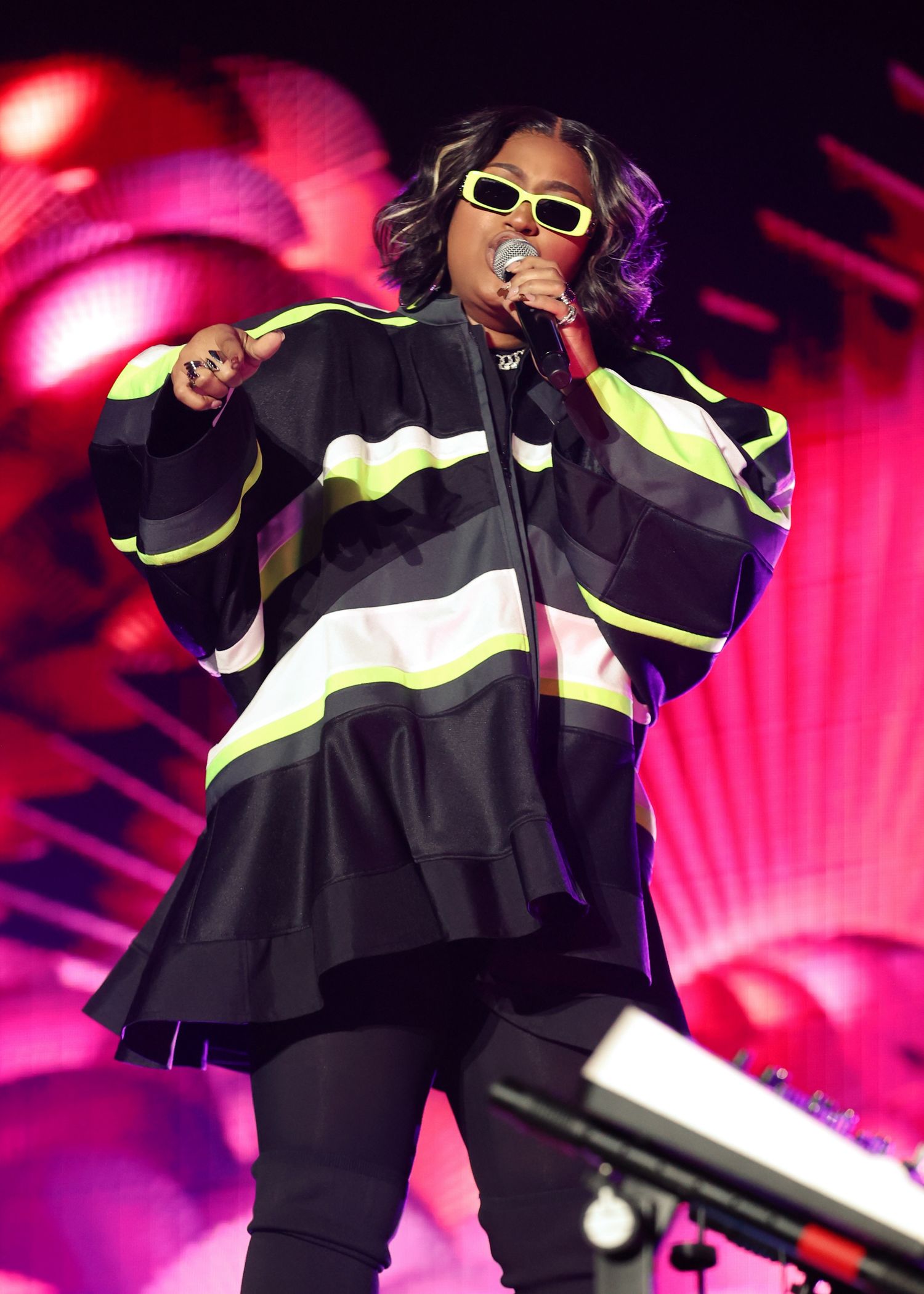 Performer in black outfit with neon accents and sunglasses dancing energetically on stage with red lighting effects.