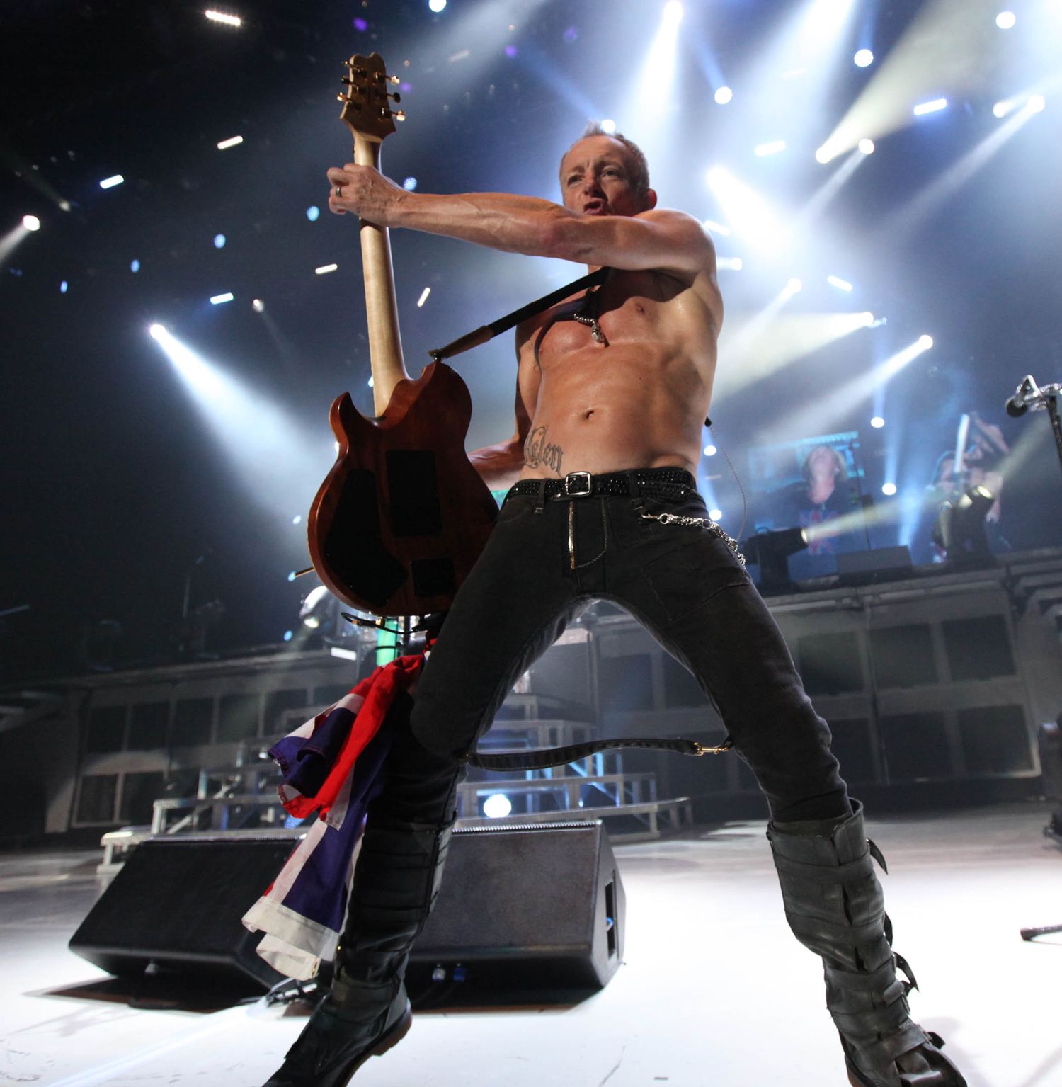 Shirtless performer holds guitar overhead during dramatic concert performance with bright stage lights.