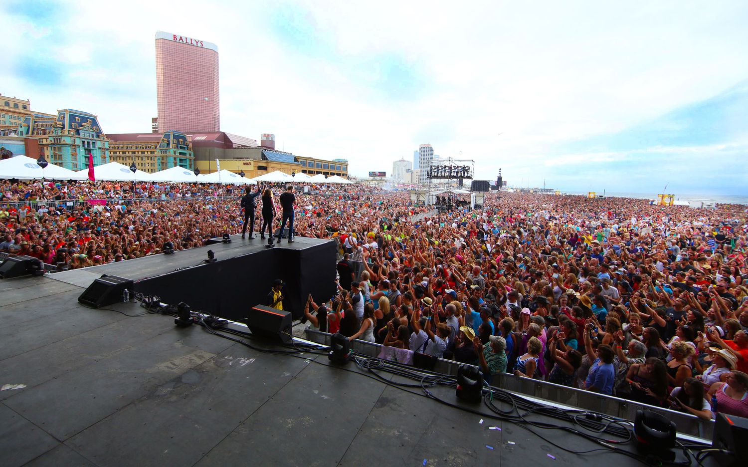 Massive crowd gathered at an outdoor concert venue with stage visible in distance.