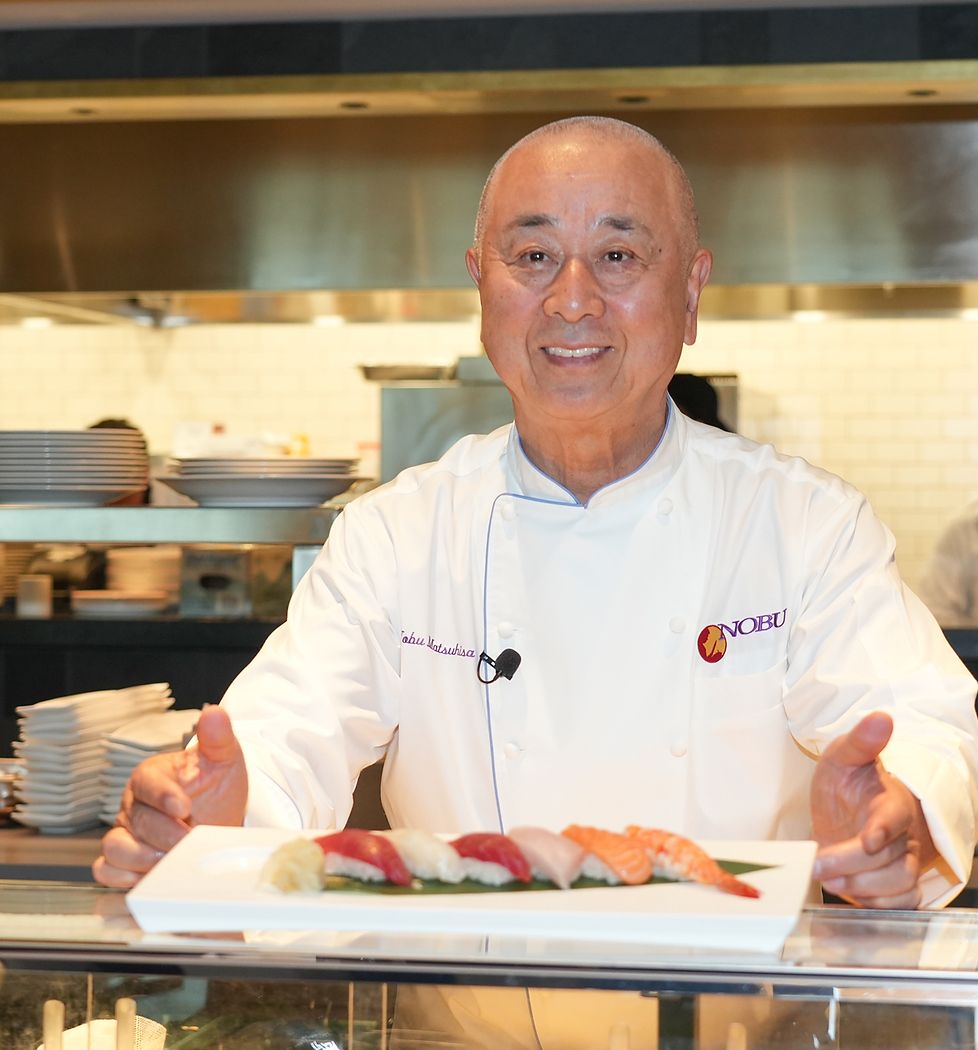 Chef in white uniform smiling while preparing sushi at a restaurant counter.