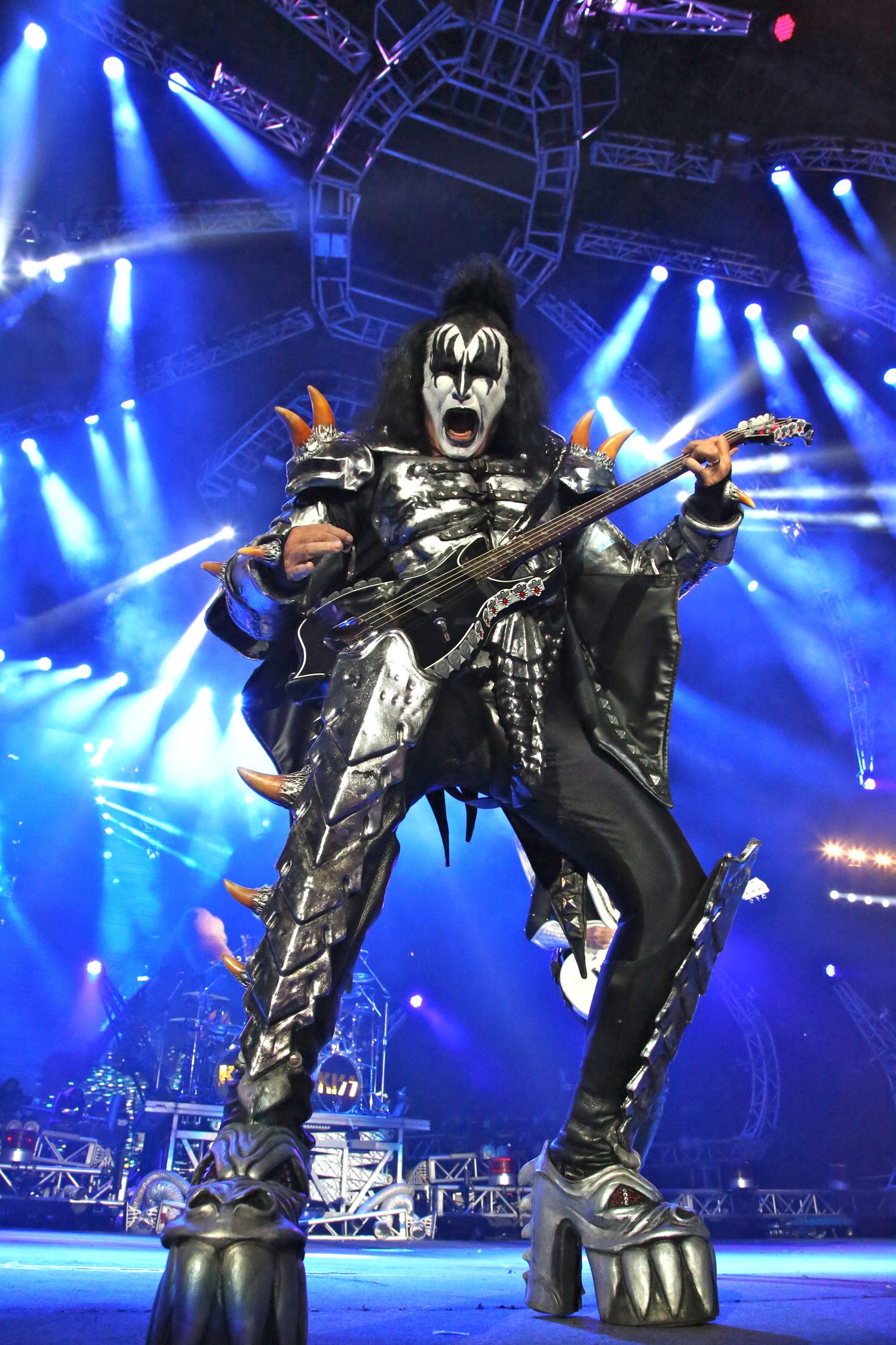 KISS band member performing in full costume and makeup with guitar against blue stage lighting.