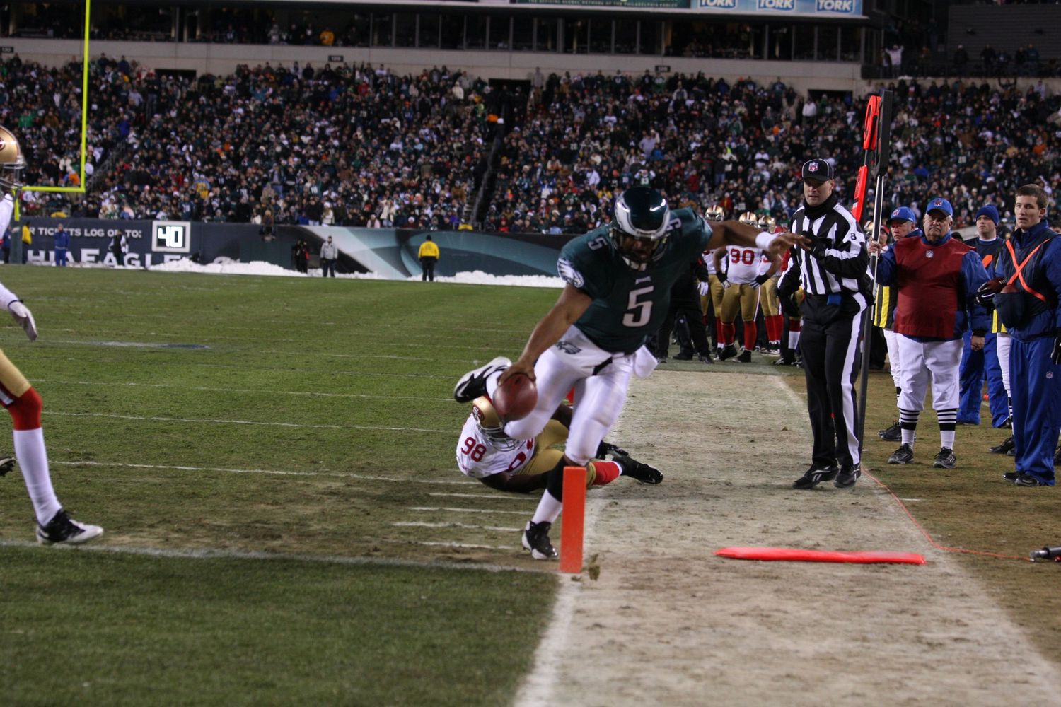 Football player leaps over defender during intense sideline play with officials watching.