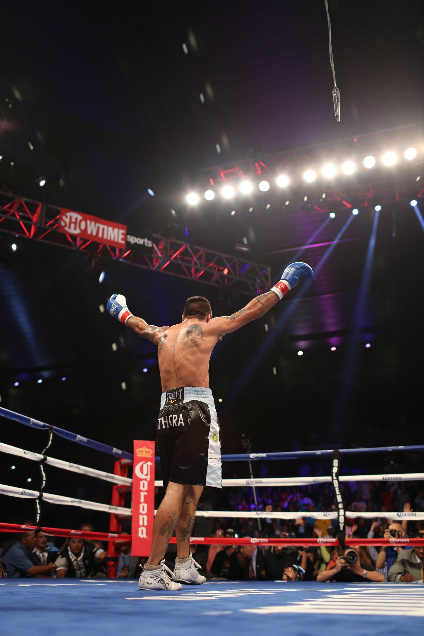 A boxer celebrates victory in the ring with arms raised under dramatic arena lighting.