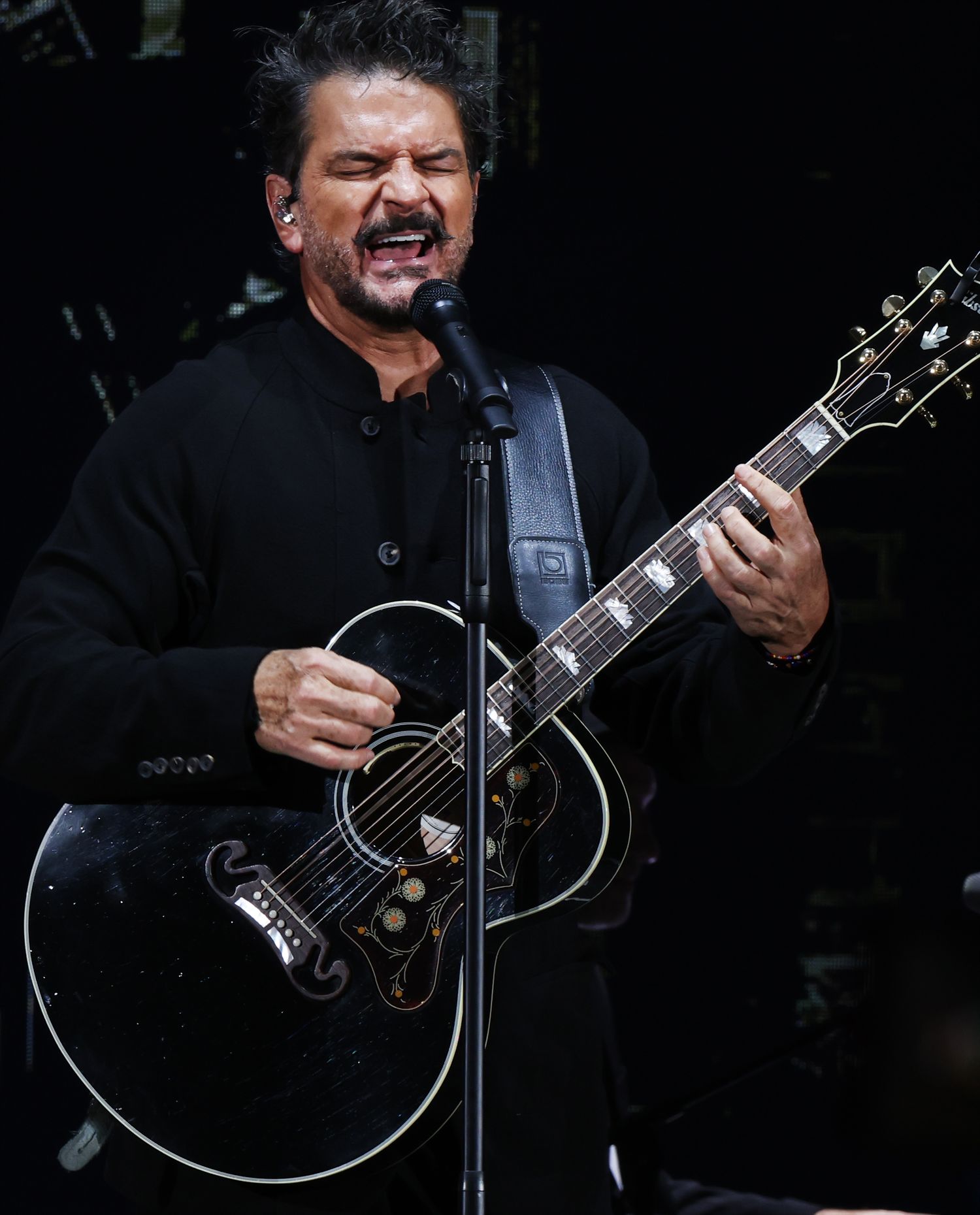 Musician in black clothing performing with acoustic guitar on dark stage.