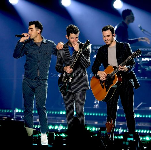 Musical group performing on stage with acoustic guitars and coordinated outfits under blue lighting.