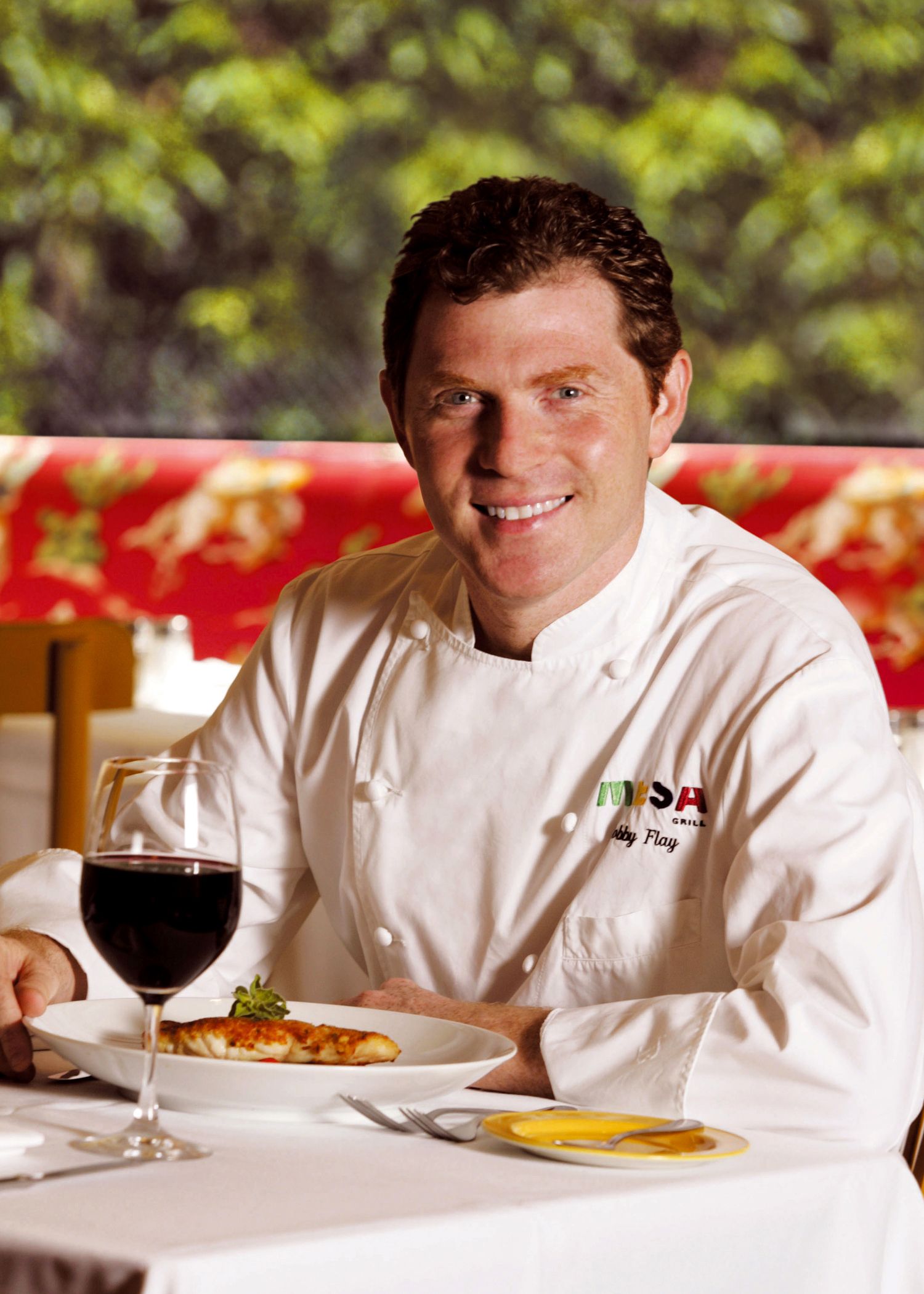 Chef in white uniform seated at dining table with wine glass and plated food.