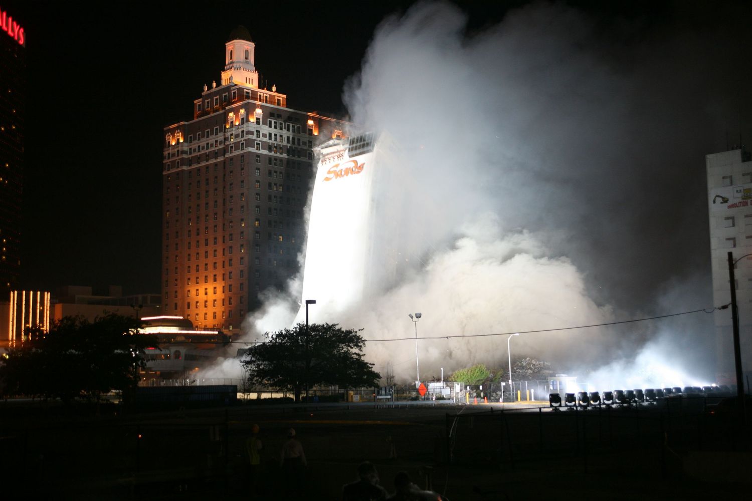 Dramatic nighttime demolition of a building with bright spotlights illuminating rising dust and debris.