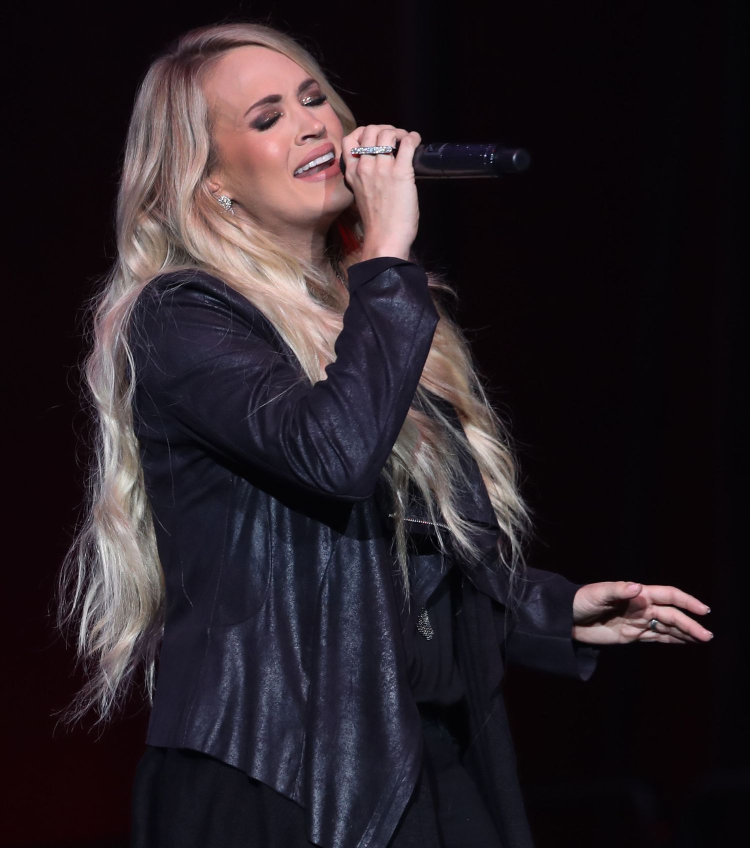 Singer in black outfit performing passionately on dark stage with dramatic lighting.