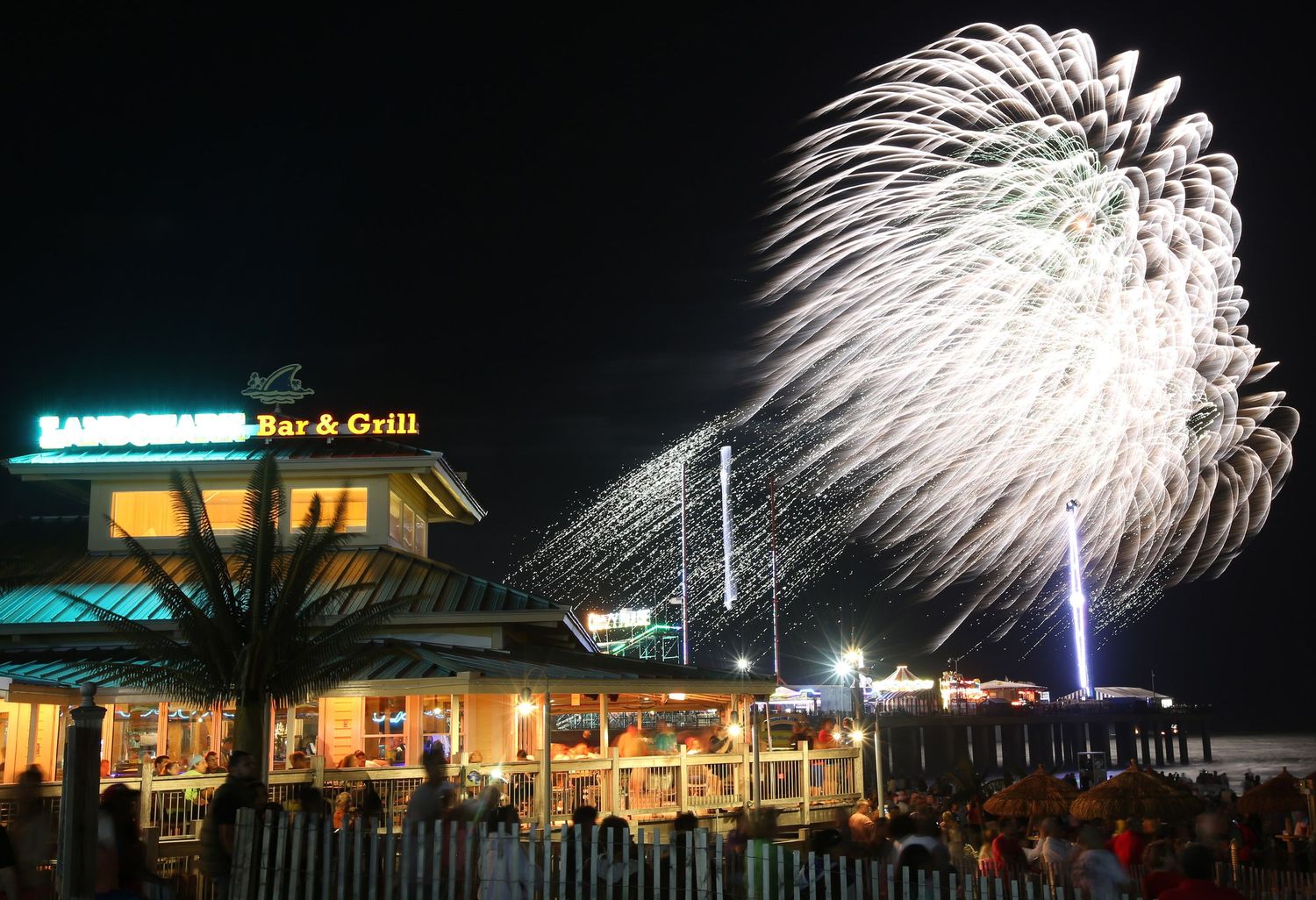Nighttime fireworks display over beachfront buildings and boardwalk.
