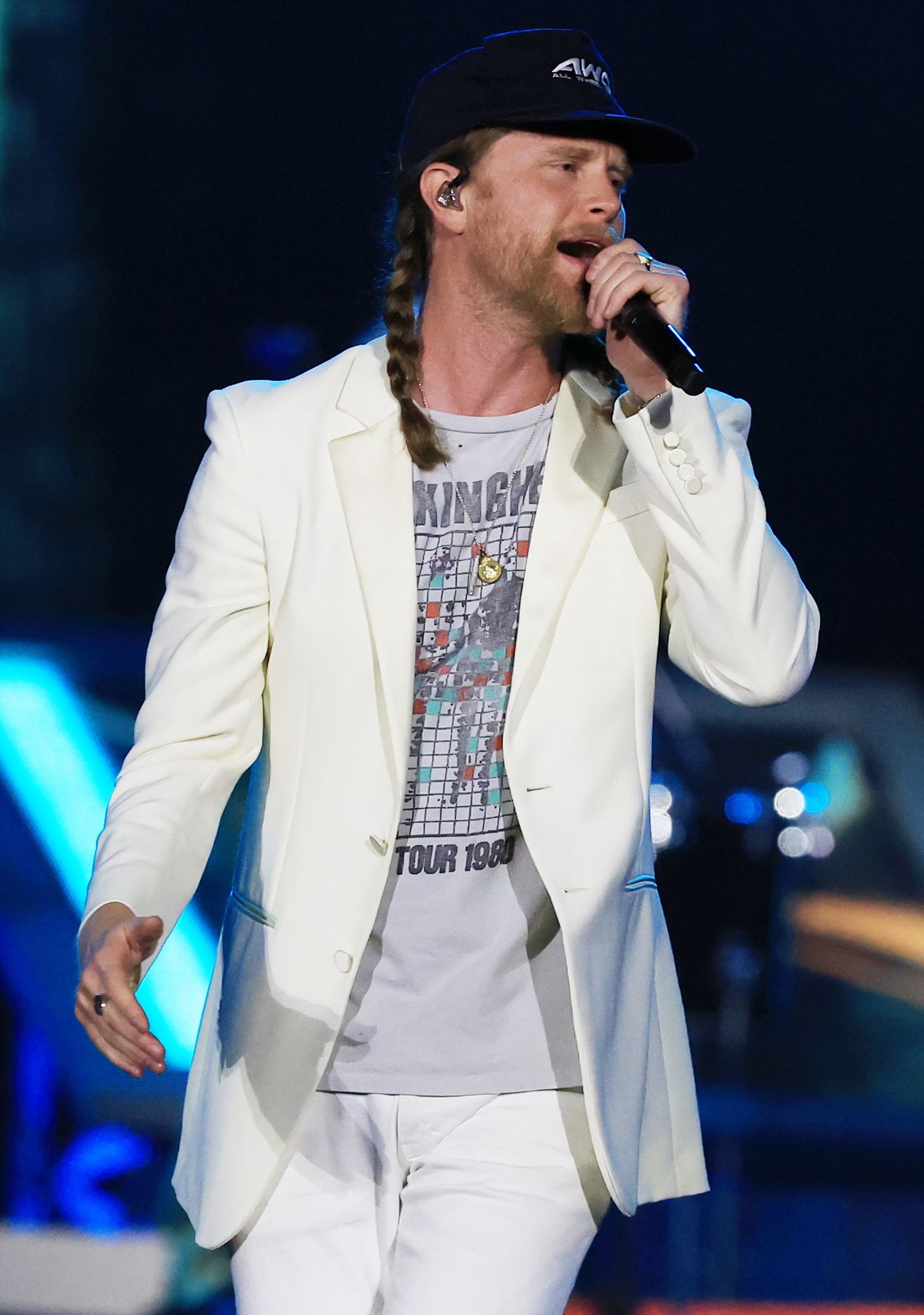 Performer in white jacket singing on stage with blue lighting effects.
